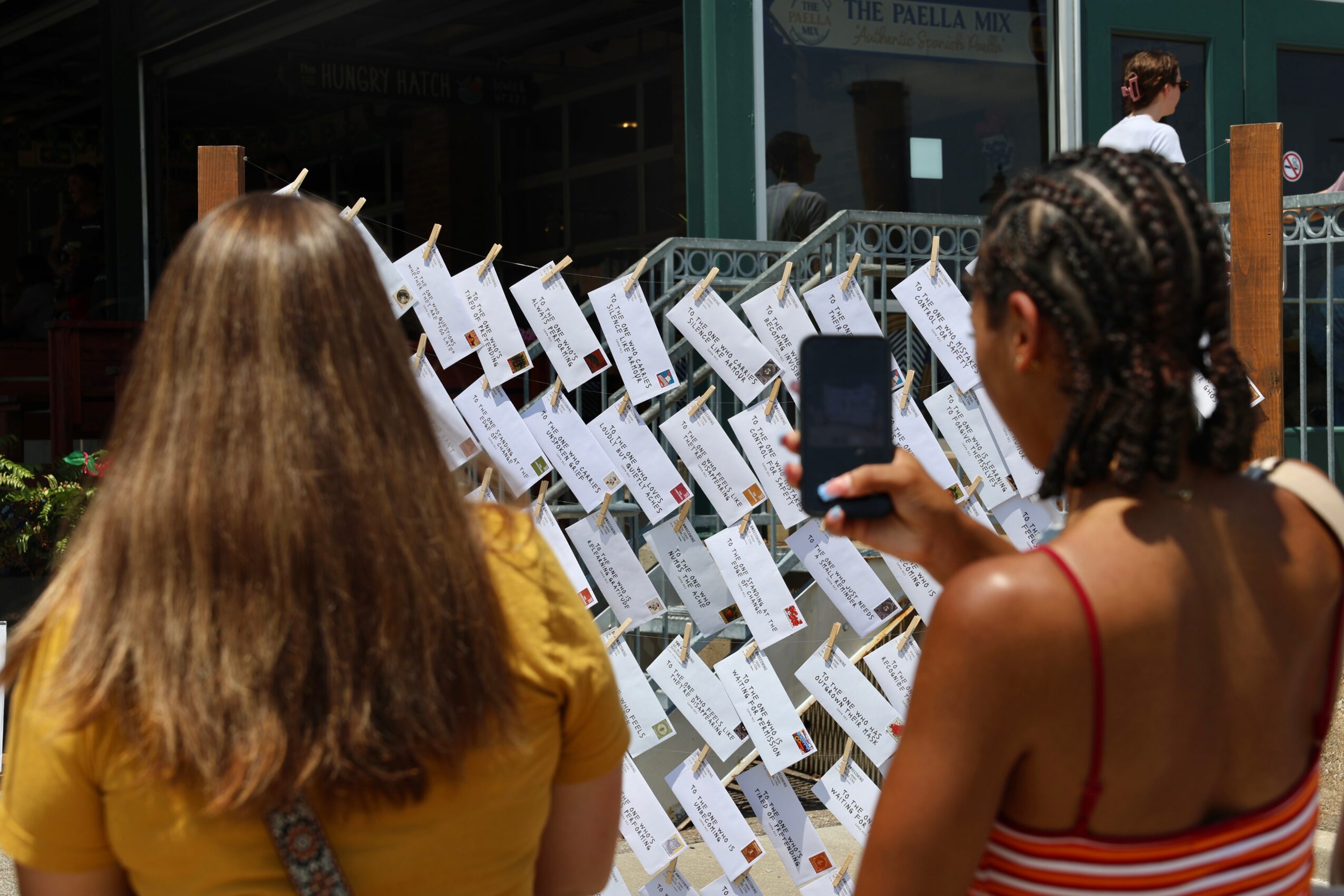 Two women stand outdoors, one with long hair in a yellow shirt, the other with braided hair in a striped top, taking a photo of a display with papers or notices. Word count: 27, confidence: 0.9