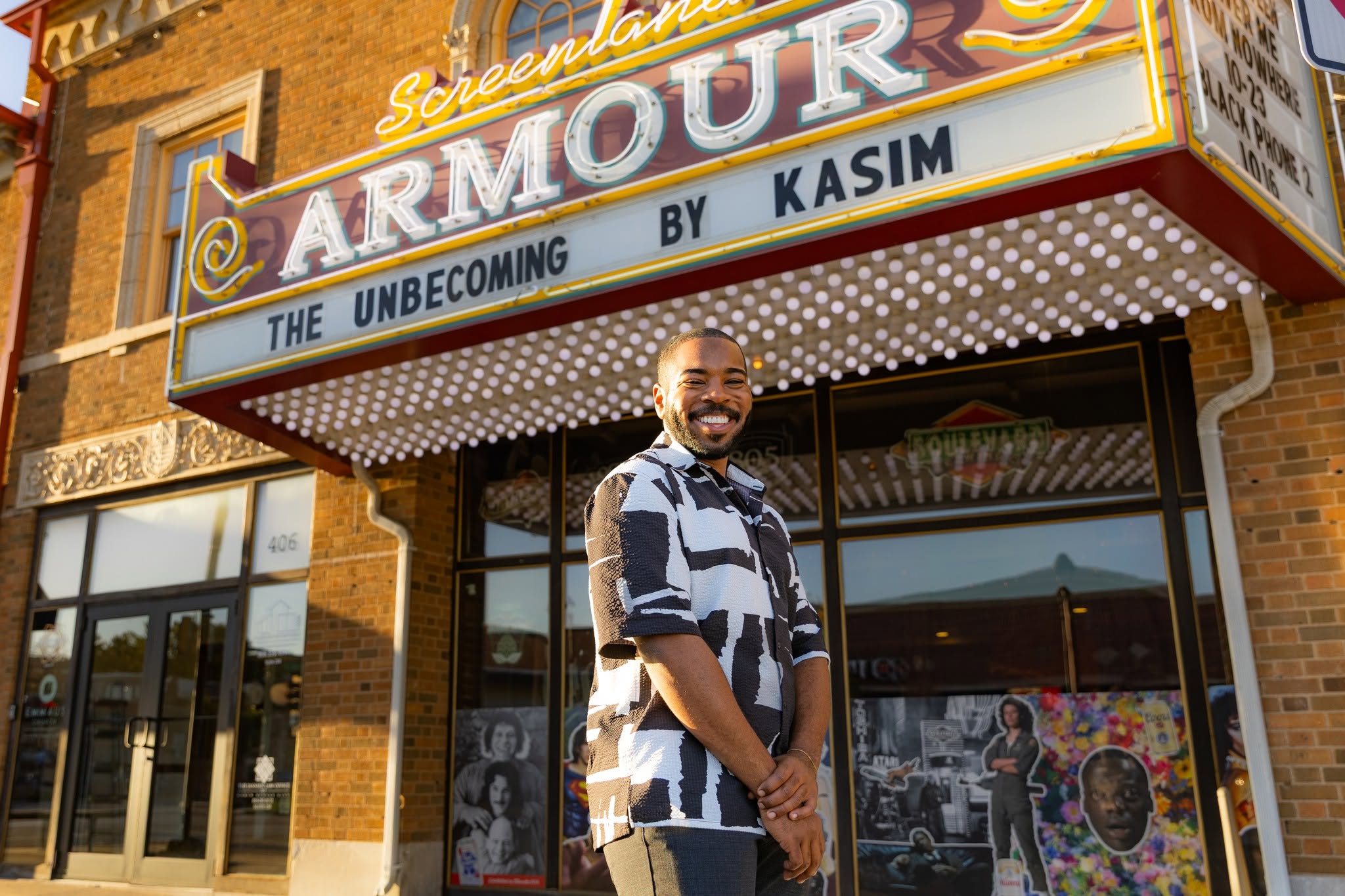 Man standing outside theater with marquee sign, smiling, brick building, glass doors, and posters visible behind him.