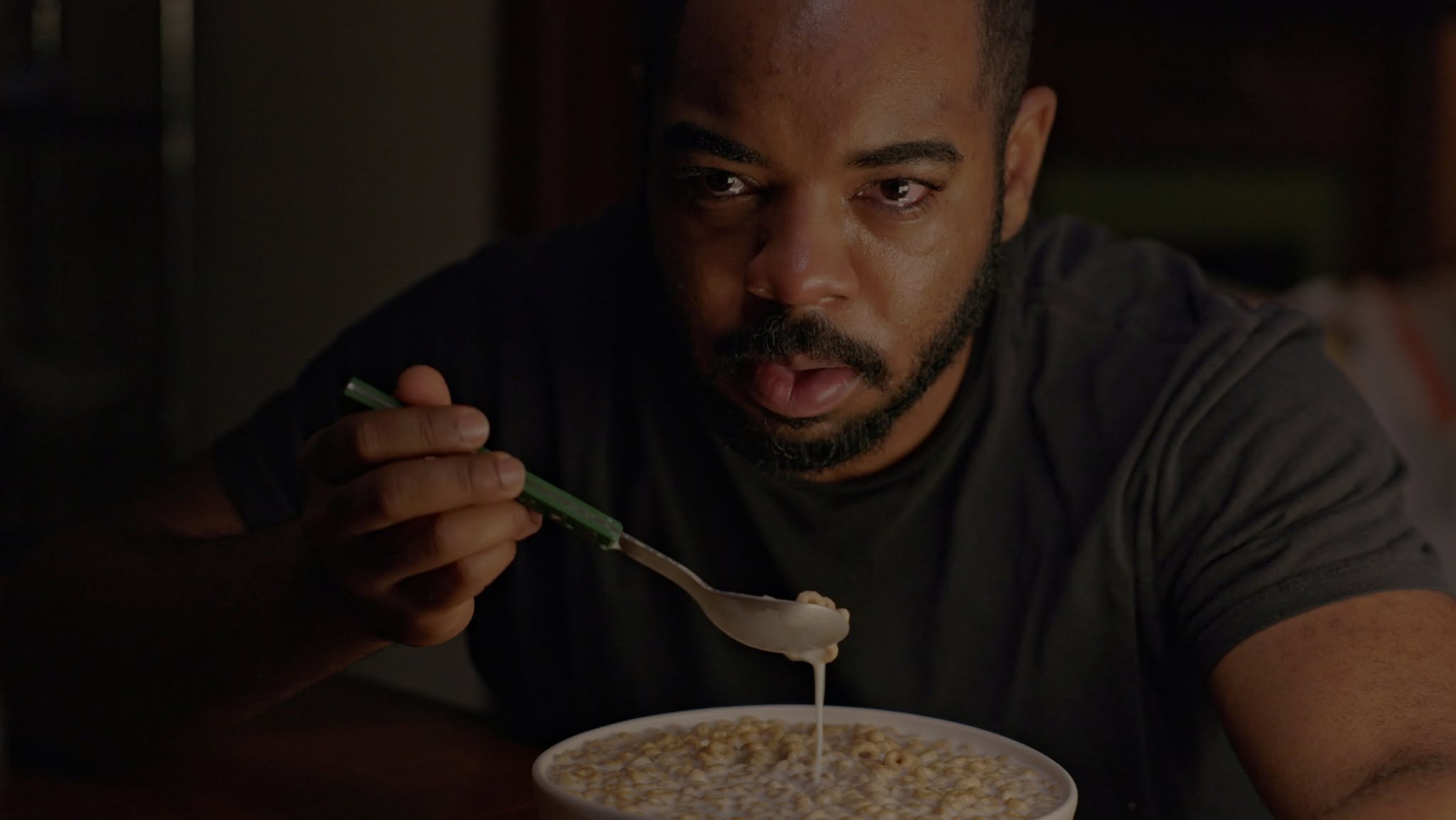 Man eating from a bowl with a spoon, dark background, focused expression.