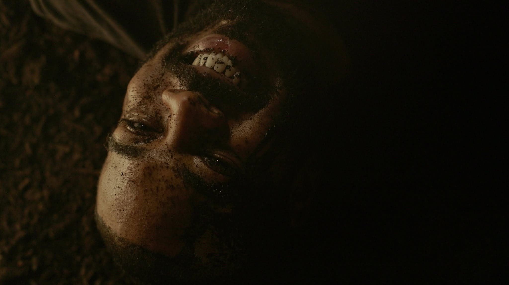 Person lying on the ground with face partially illuminated, dark background, face covered in dirt or makeup.