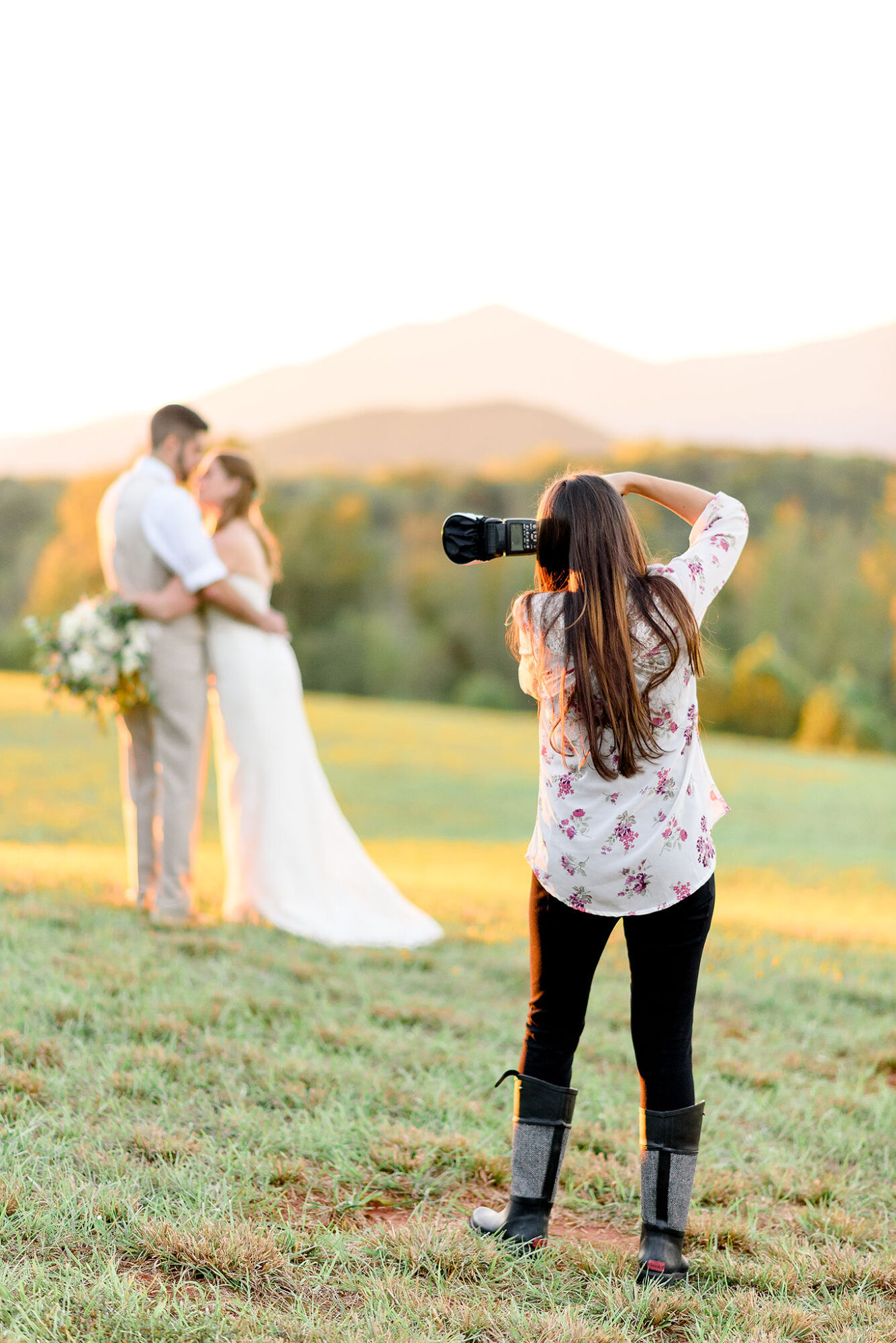 Photographer taking picture of a couple outdoors during sunset, with mountains in the background, woman holding a bouquet, man in white shirt.