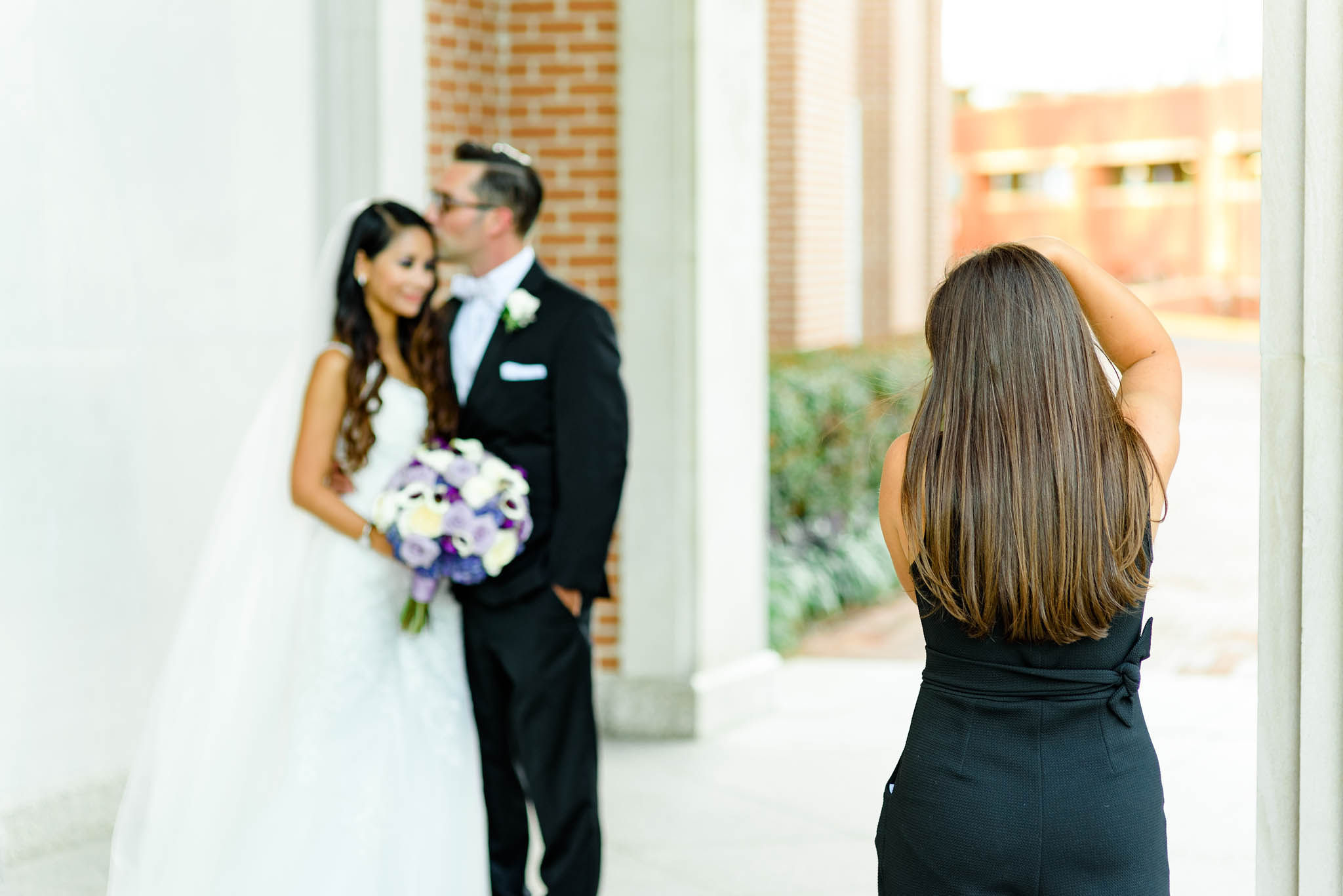 Person with long brown hair taking a photo of a bride and groom outside, bride holding a bouquet, background with brick and greenery.