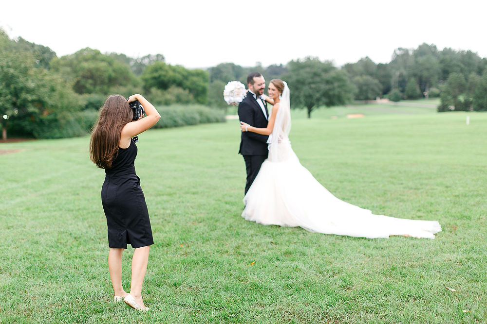 A woman takes a photo of a bride and groom standing on a grassy field with trees in the background.
