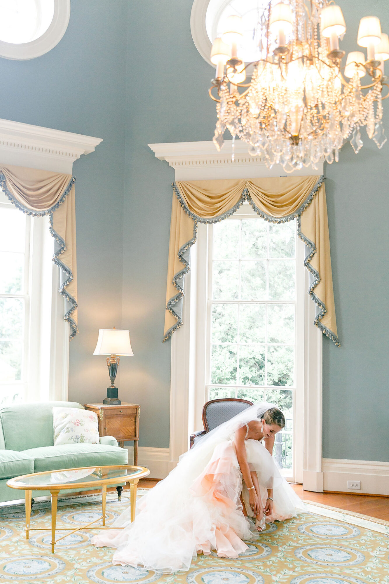 Bride in a wedding dress kneeling in a luxurious room with large windows, chandelier, and elegant curtains.