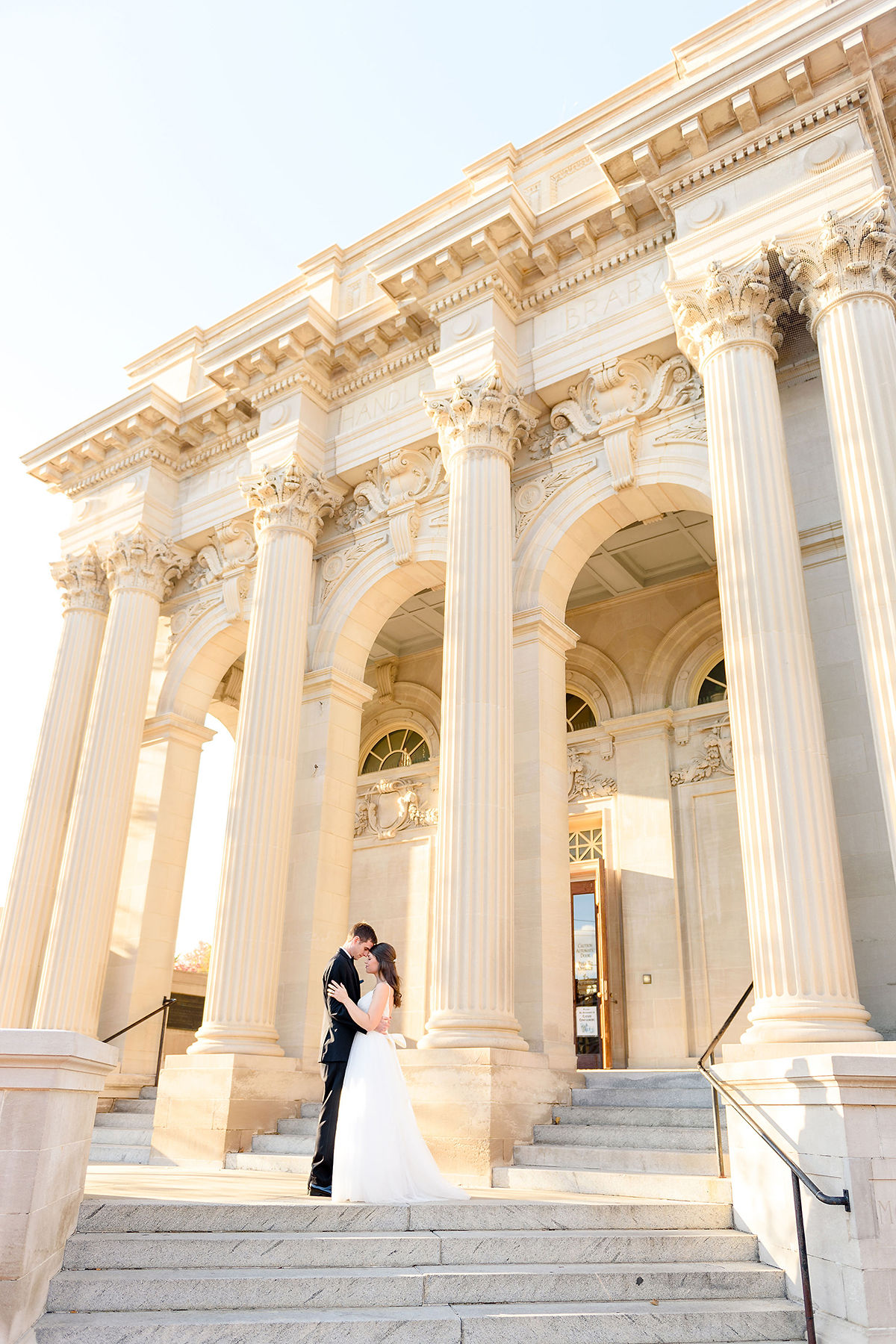 A couple in wedding attire stands on steps in front of a grand classical building with tall columns.