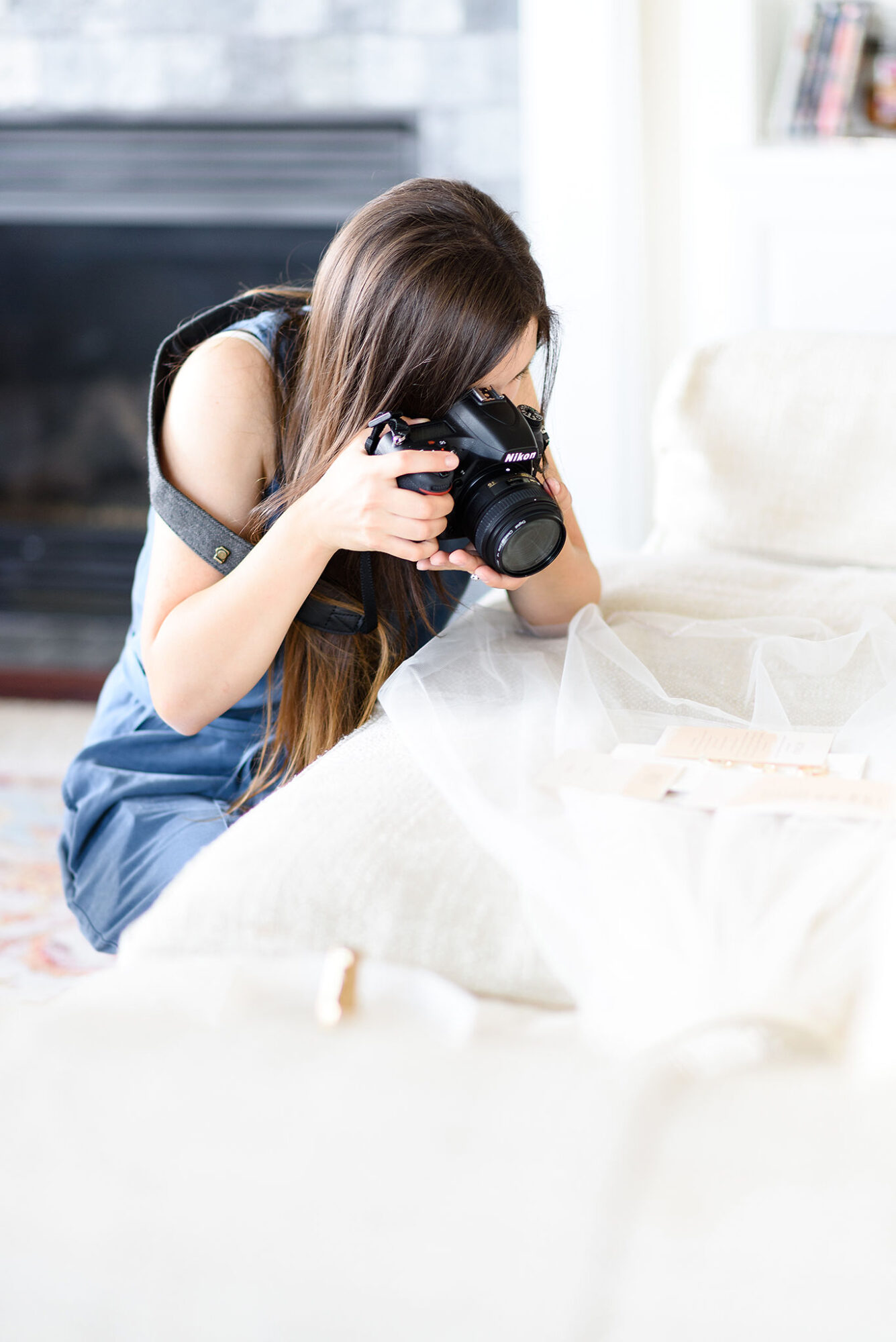 Young woman with long brown hair holding a camera, looking through the viewfinder, sitting indoors near a white surface.