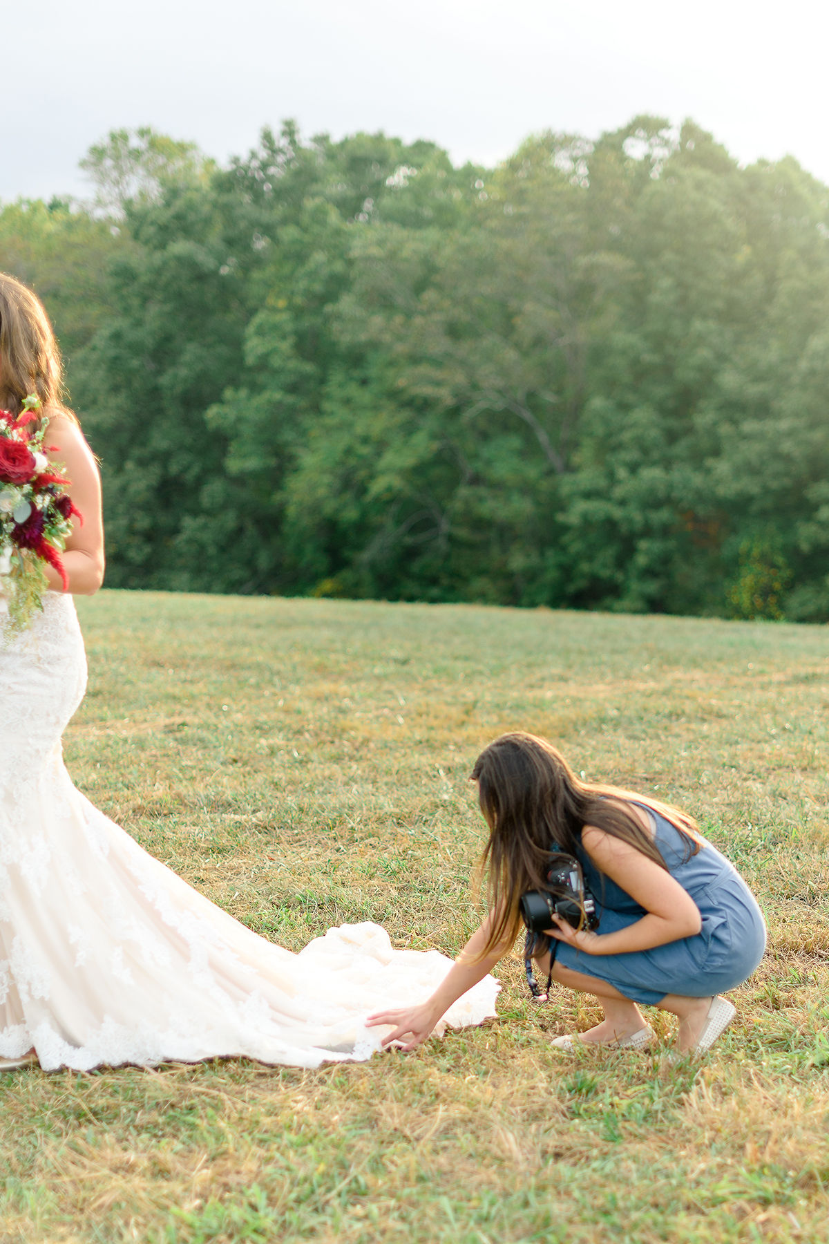 A girl in a blue dress crouches on grass, touching the train of a wedding dress outdoors with trees in background.