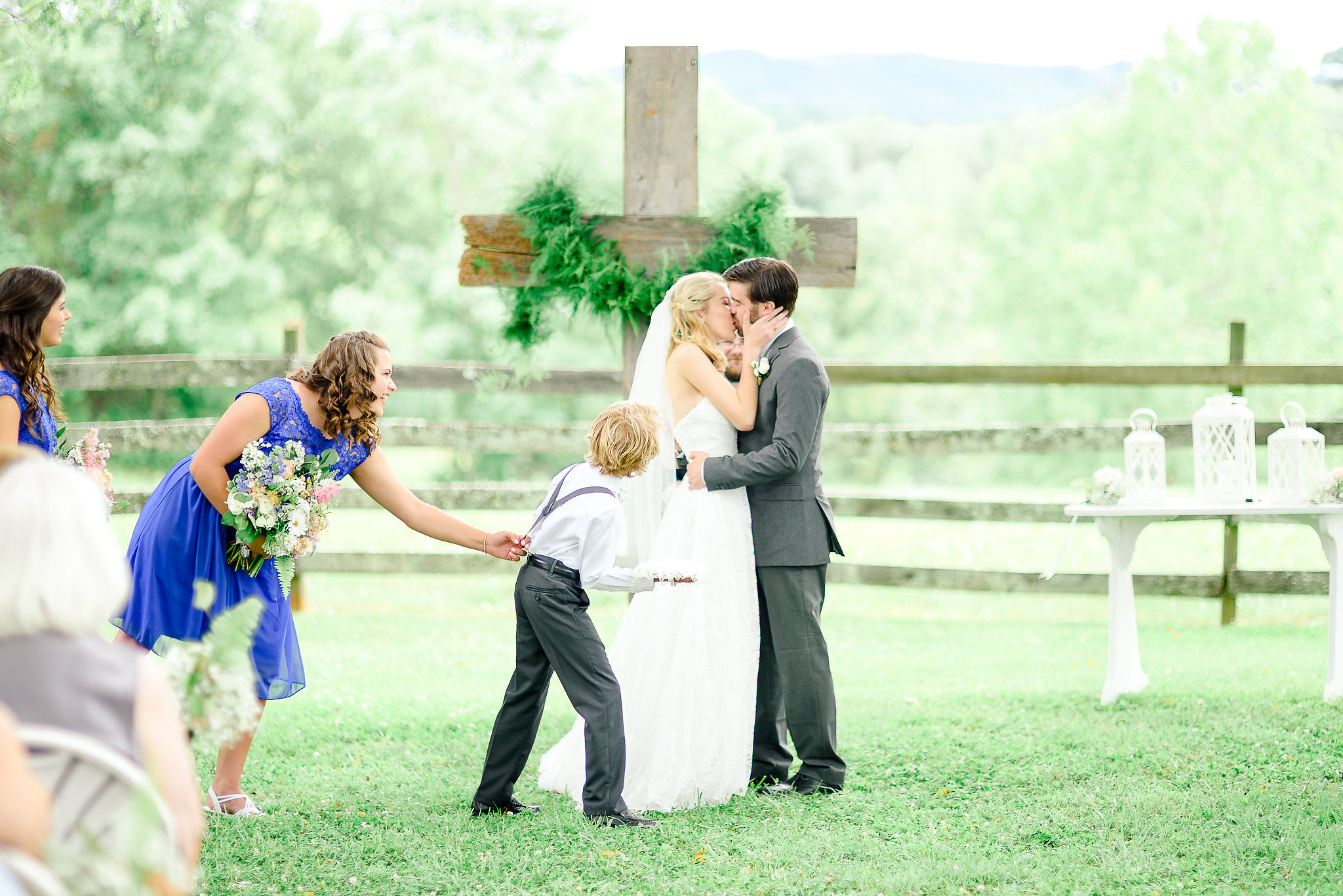 A wedding ceremony outdoors with a couple kissing, surrounded by children and guests, with a cross and greenery in the background.