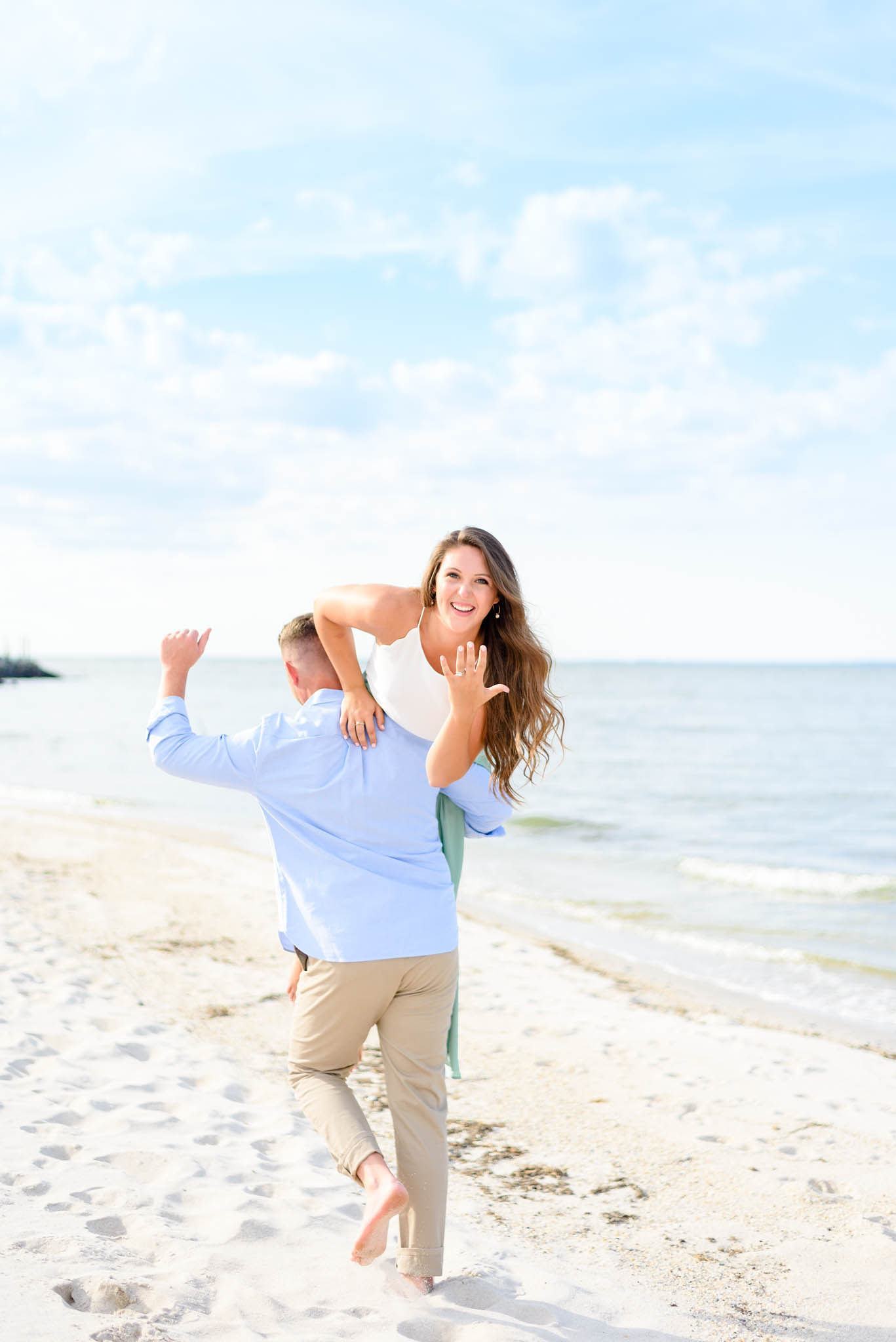 A woman is carried on a man's shoulders on a beach, smiling, with the ocean and sky in the background.