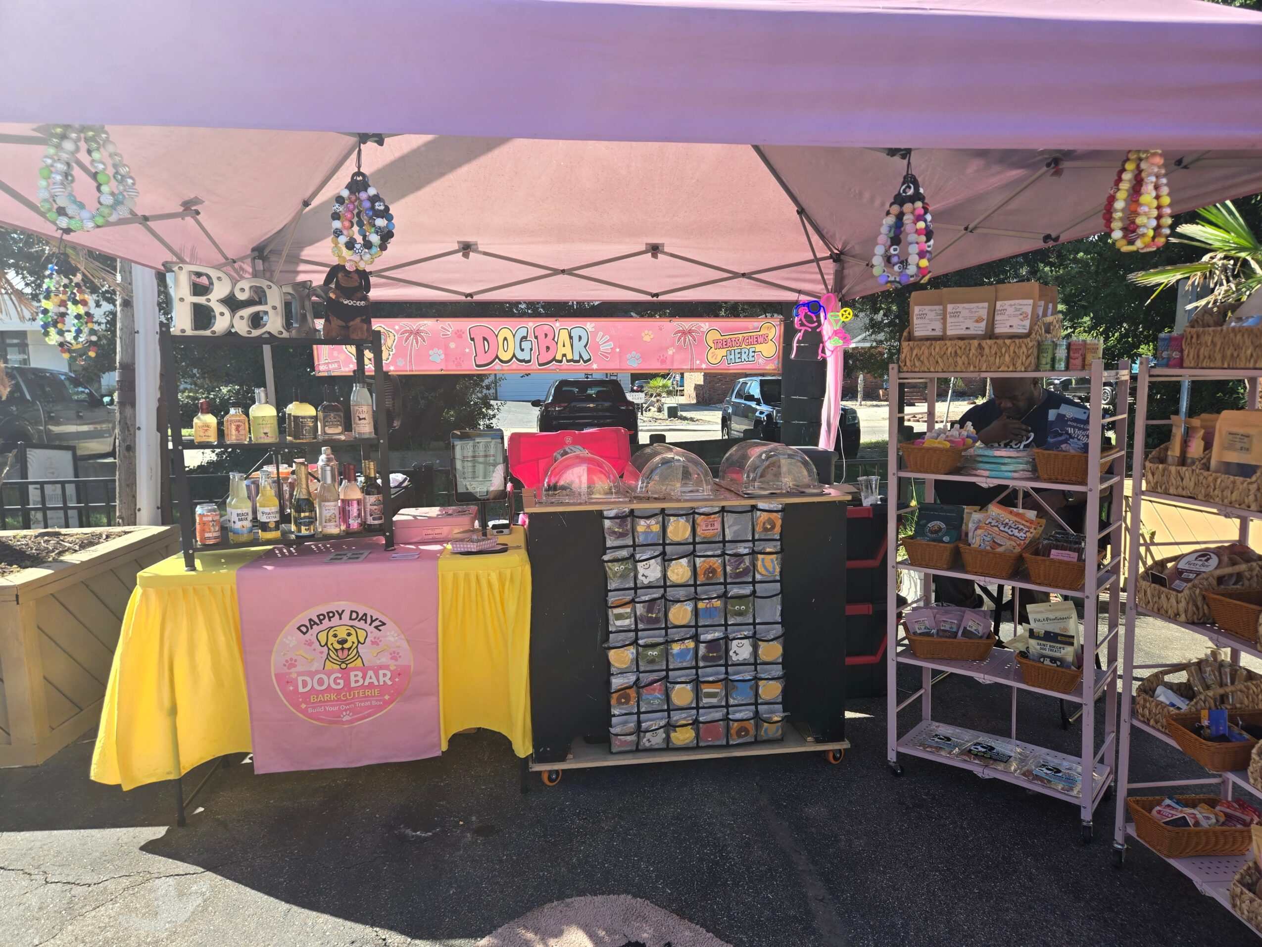 Food and drink stand with a pink sign, yellow tablecloth, and shelves of items, under a pink canopy.