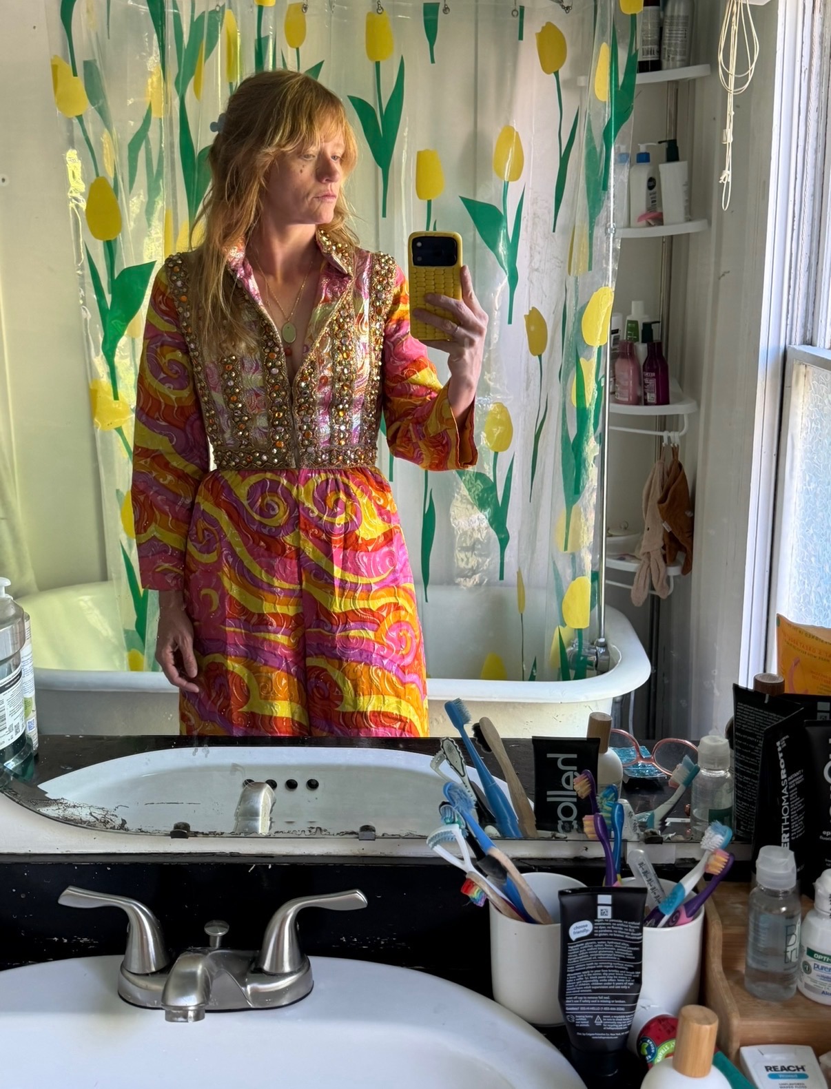 Woman with long hair taking a mirror selfie in a bathroom with floral shower curtain, wearing a colorful patterned dress.