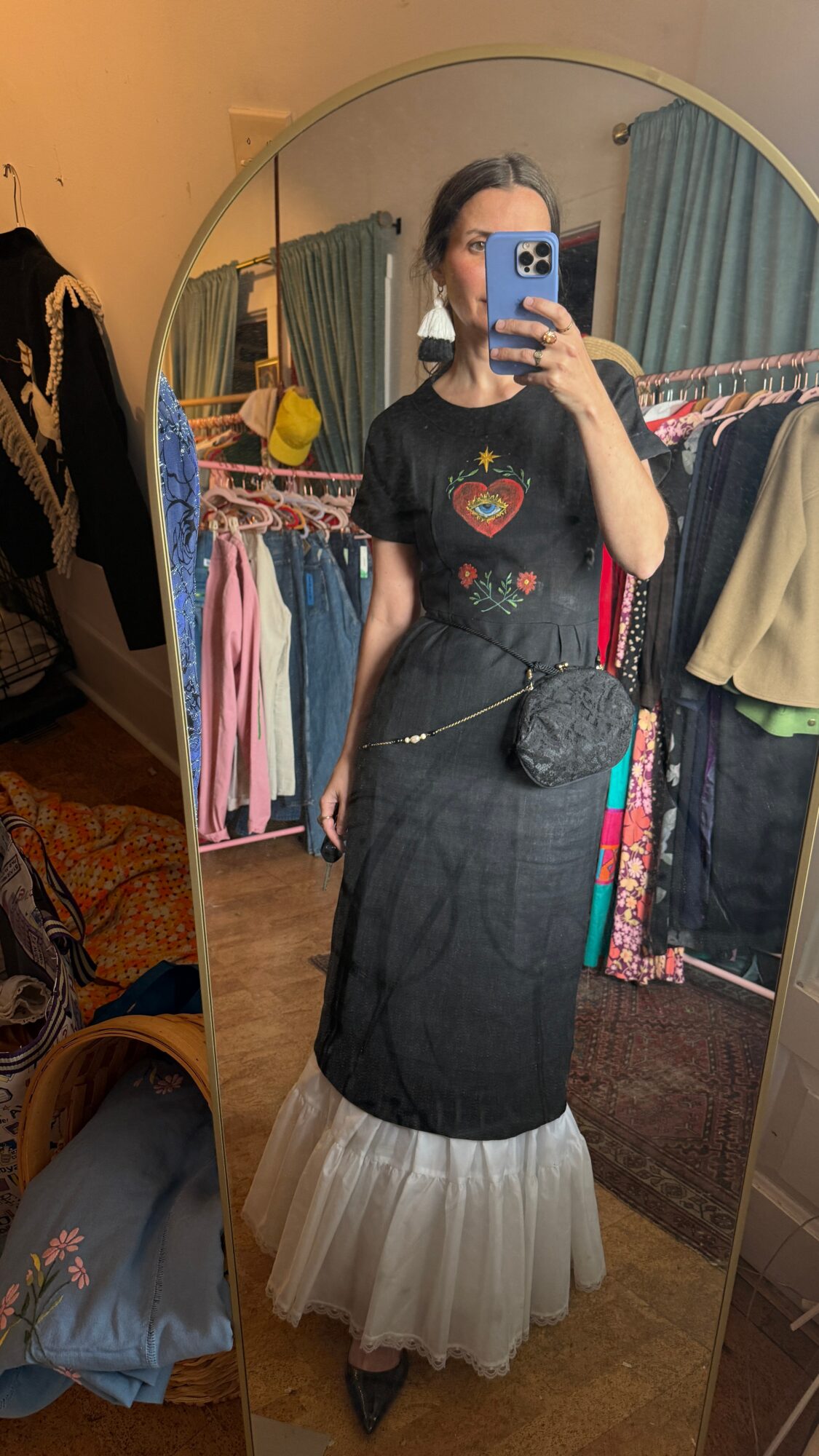 Person taking mirror selfie wearing a black top and layered skirt, in a room with clothes and a mirror.