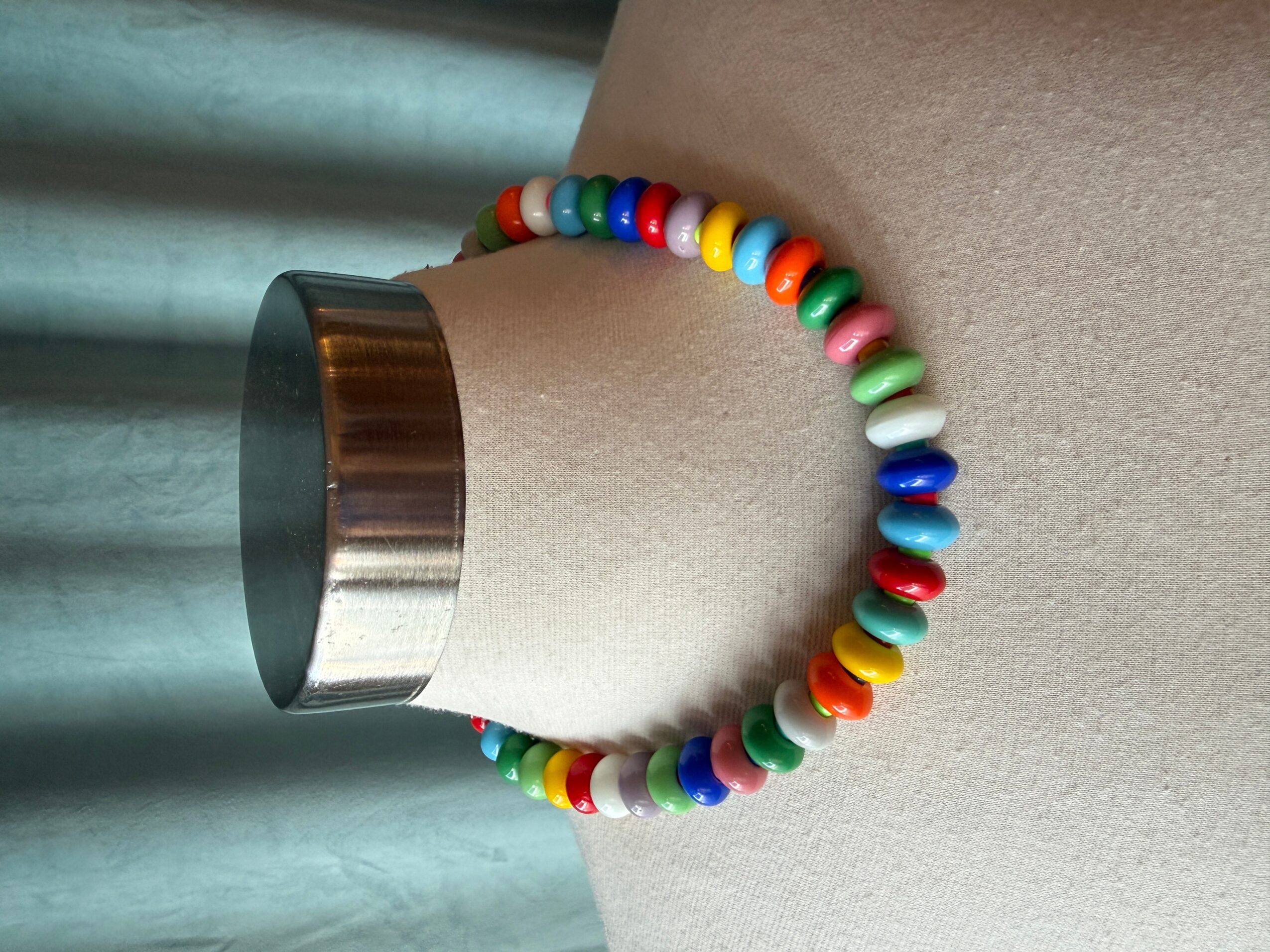 Colorful beaded bracelet on a person's wrist, with a metallic cylindrical object nearby, against a neutral background.
