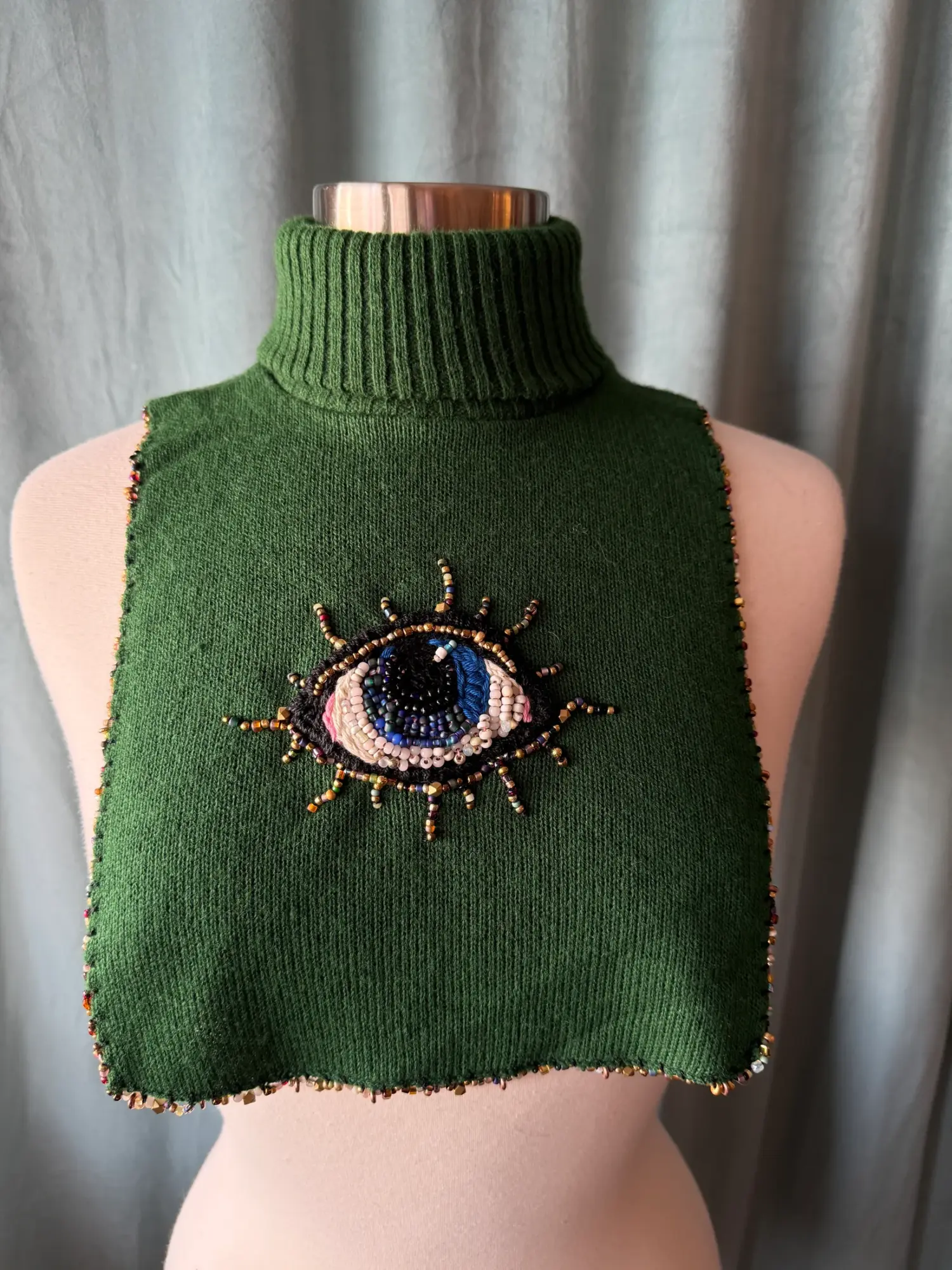 Green sleeveless turtleneck sweater with an embroidered eye design on the front, displayed on a mannequin against a curtain background.