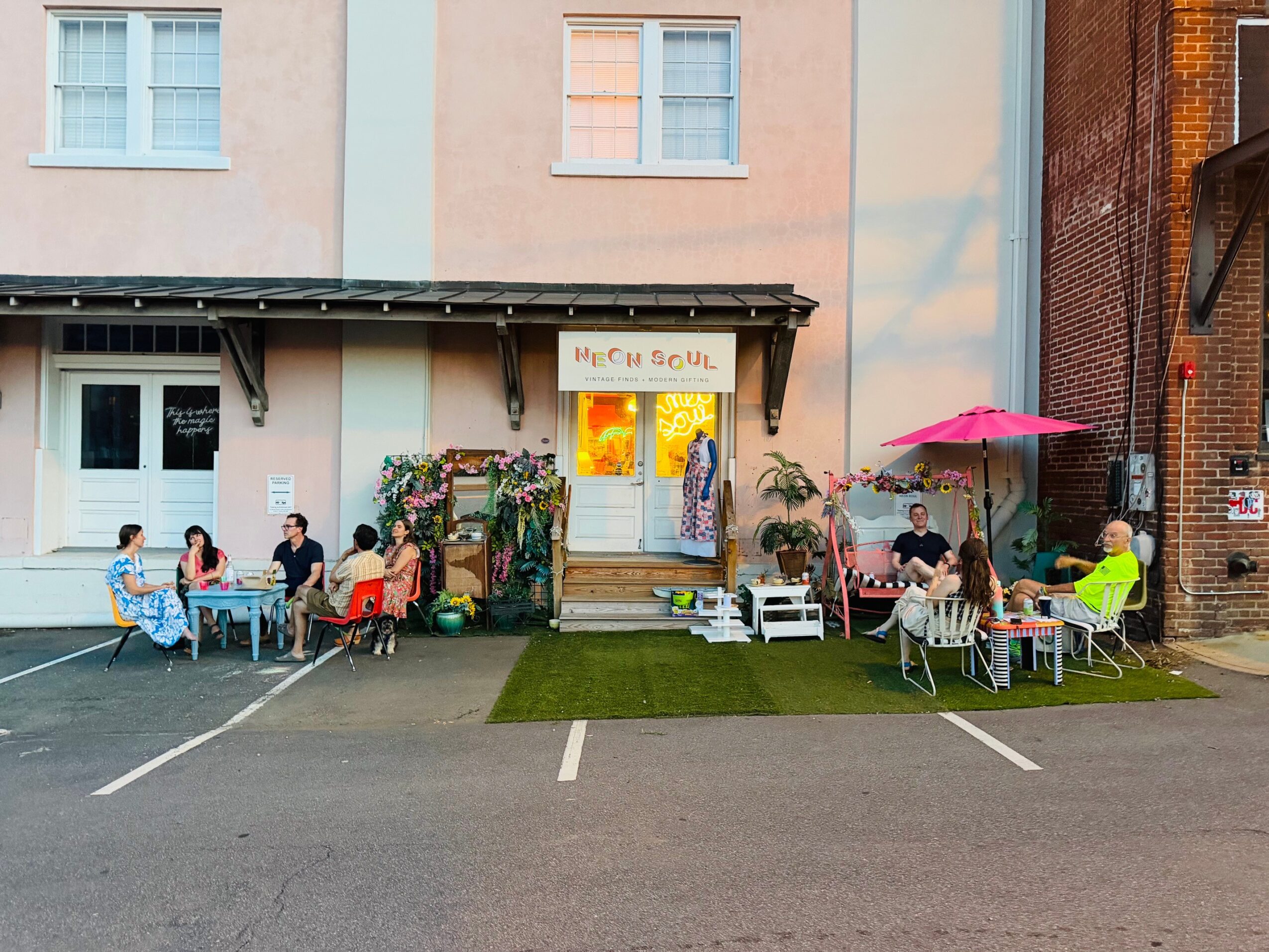 People sitting at outdoor tables with umbrellas outside a building with a sign, some greenery, and a parking lot.