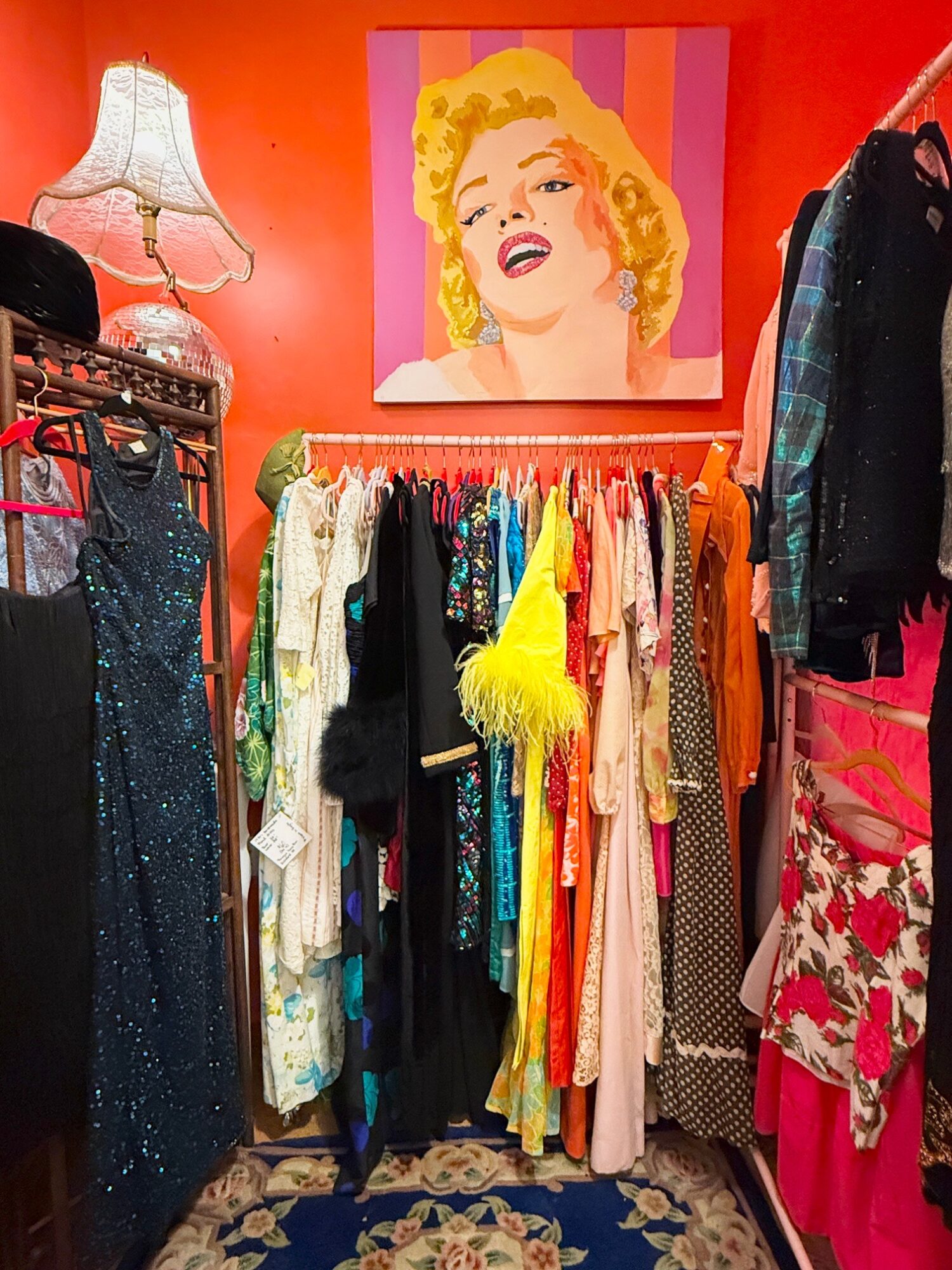 Clothing rack with colorful dresses and garments, pink wall, portrait of a woman with blonde hair, lamp, and patterned rug.