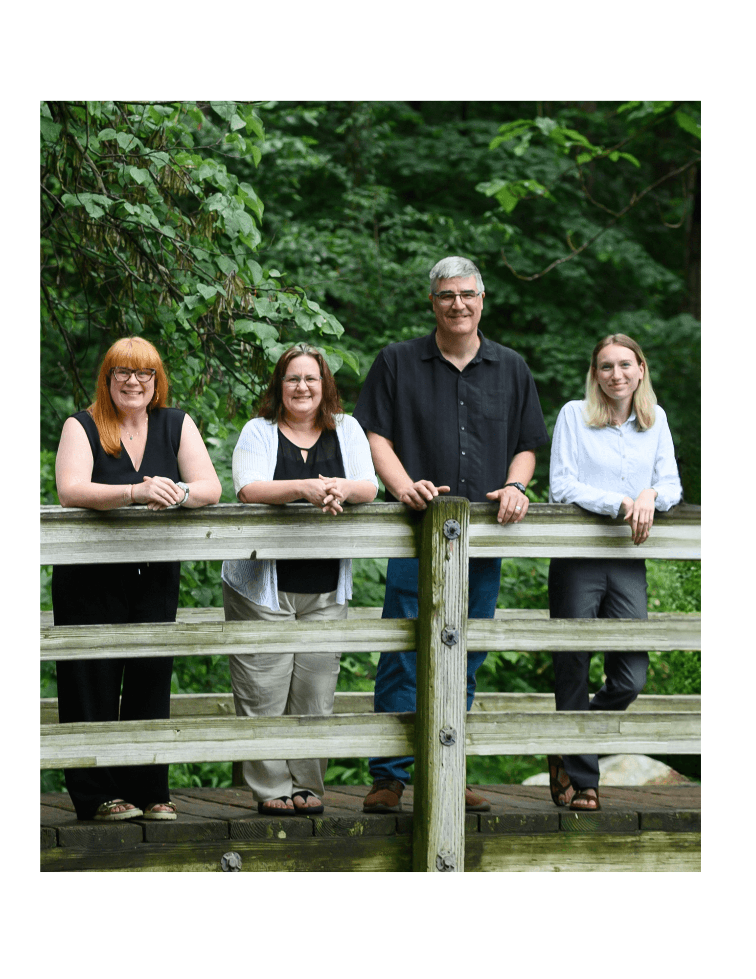 Four people stand behind a wooden railing outdoors with green trees in the background.