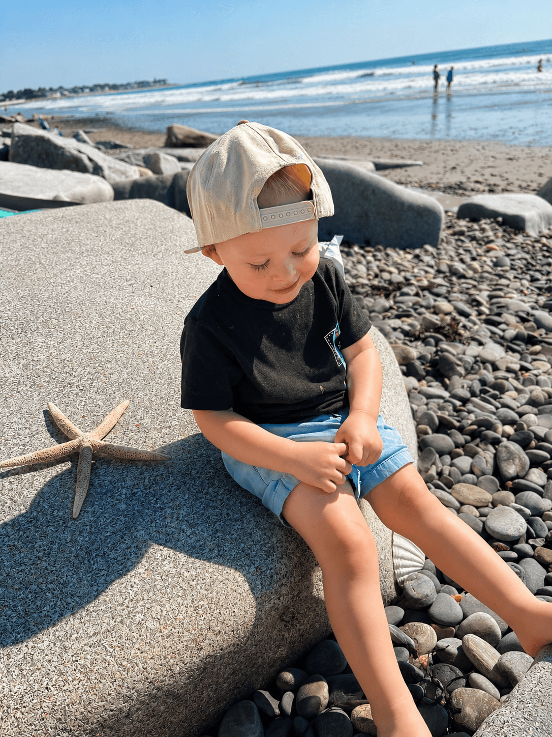 Young child sitting on rocks at the beach, wearing a beige cap and black shirt, with a starfish nearby, ocean in background.