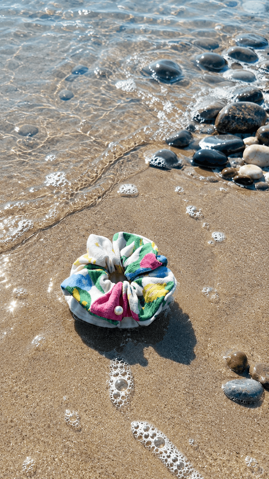 Colorful scrunchie on sandy beach with small waves and rocks, sunlight reflecting on water, shadow cast on sand.