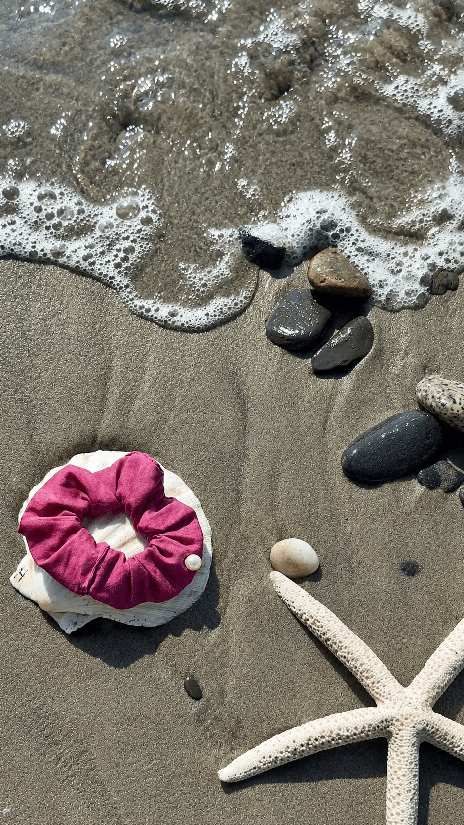 Beach scene with rocks, a starfish, a pink scrunchie, and foamy waves on sand.