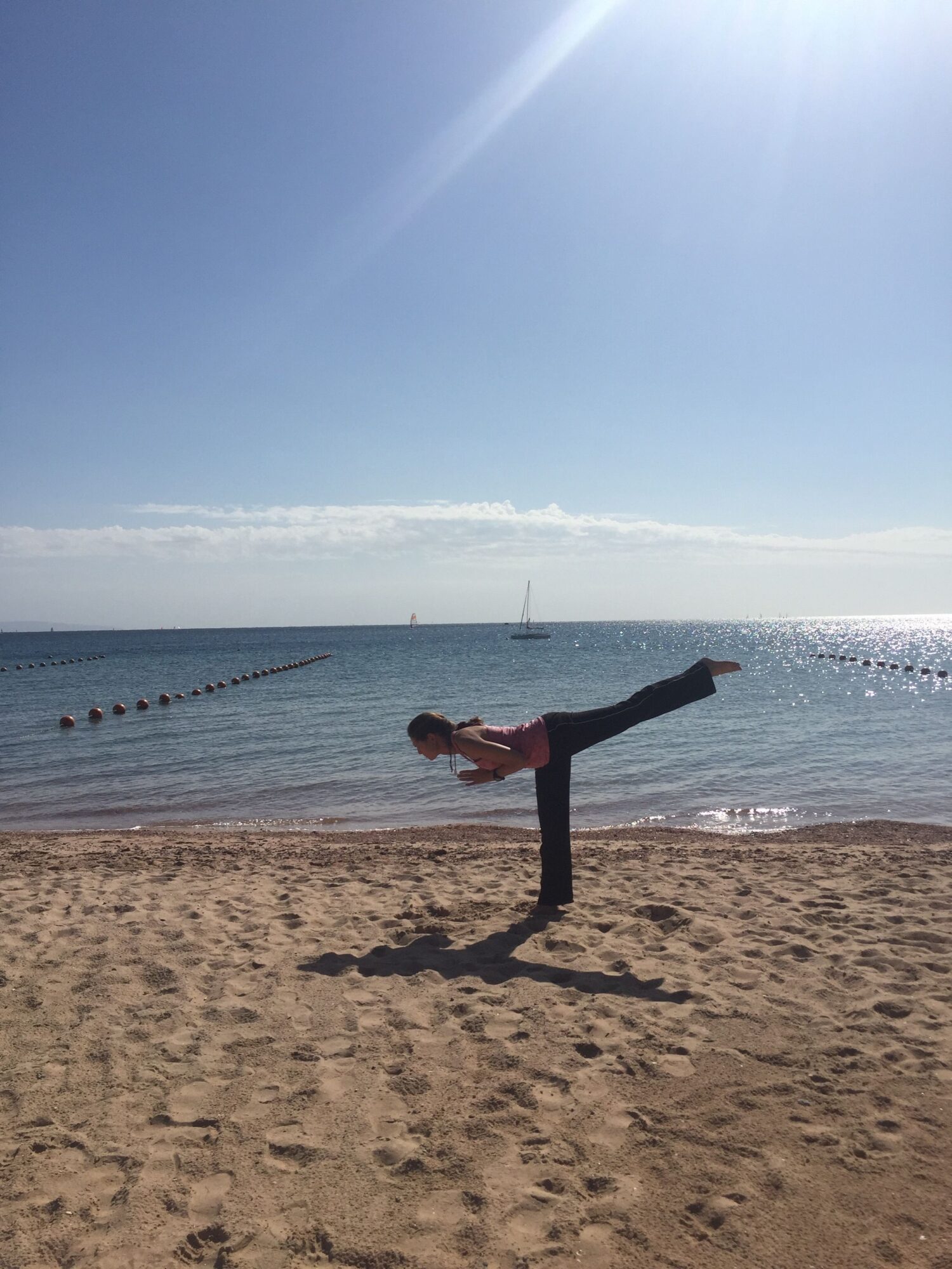 Person doing a handstand on sandy beach near water, with a boat and distant land in background.