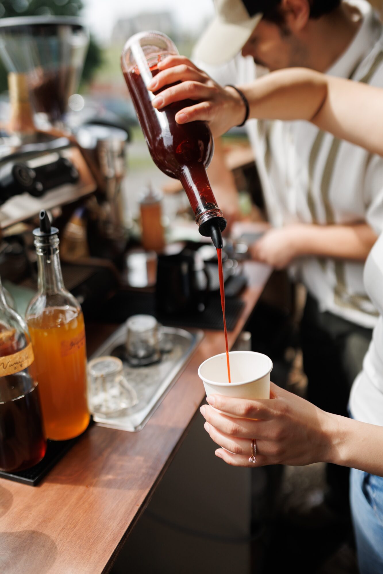 Person pouring a red drink from a bottle into a cup at a bar, with bar tools and bottles nearby.