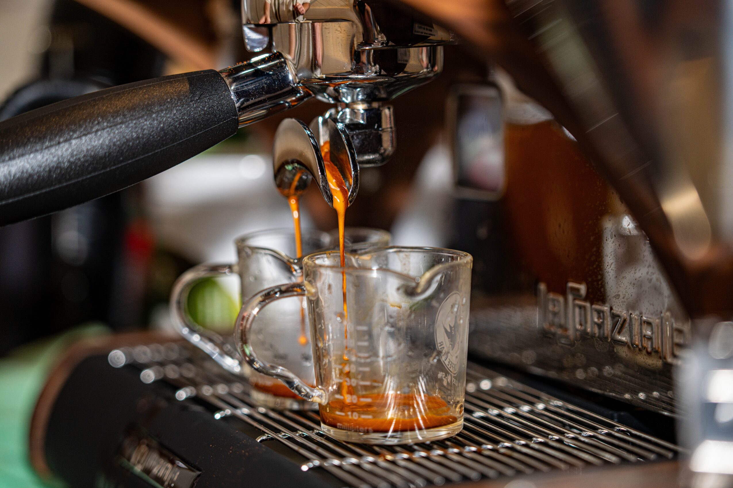 Espresso coffee flows from a machine into a glass cup, with a blurred background of coffee equipment.