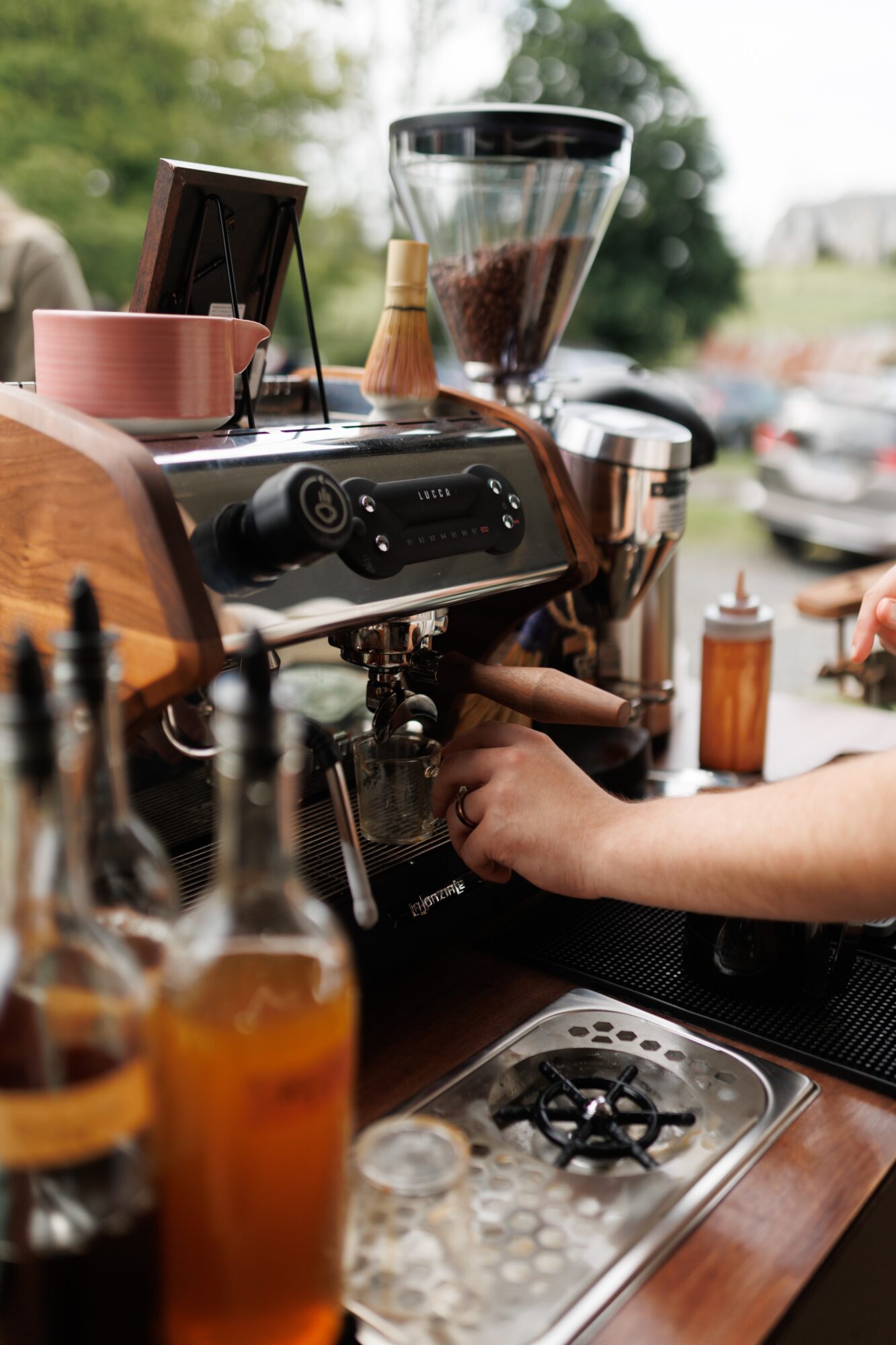 Barista's hand preparing coffee behind a coffee machine with a blender, cups, and bottles on a wooden counter outdoors.