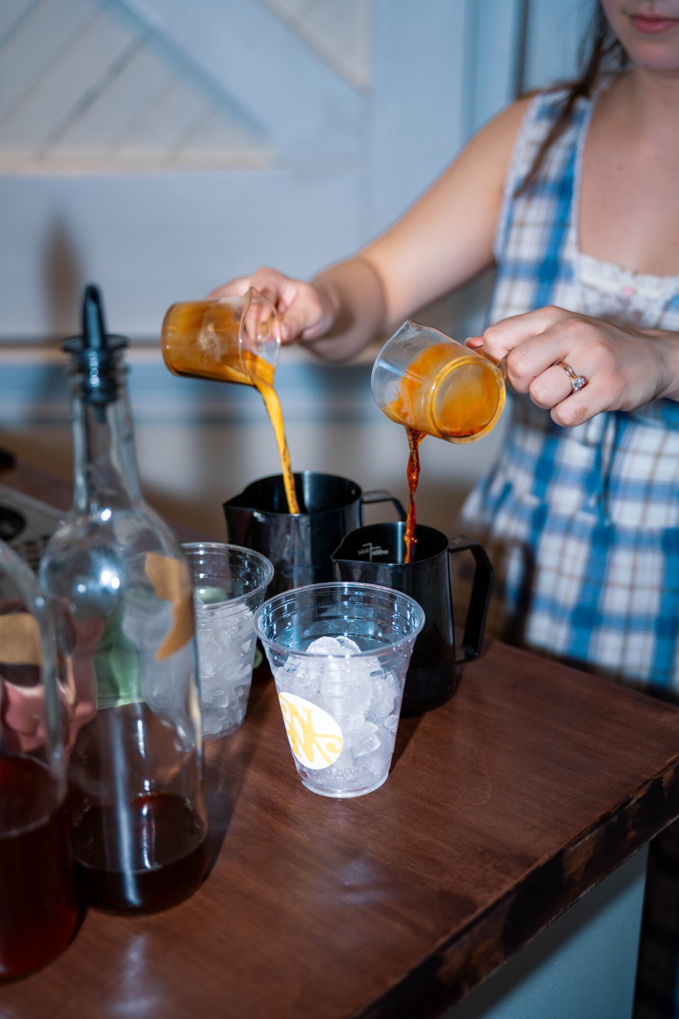 Person pouring a drink from a small glass into a plastic cup, with bottles and glasses on a wooden table.