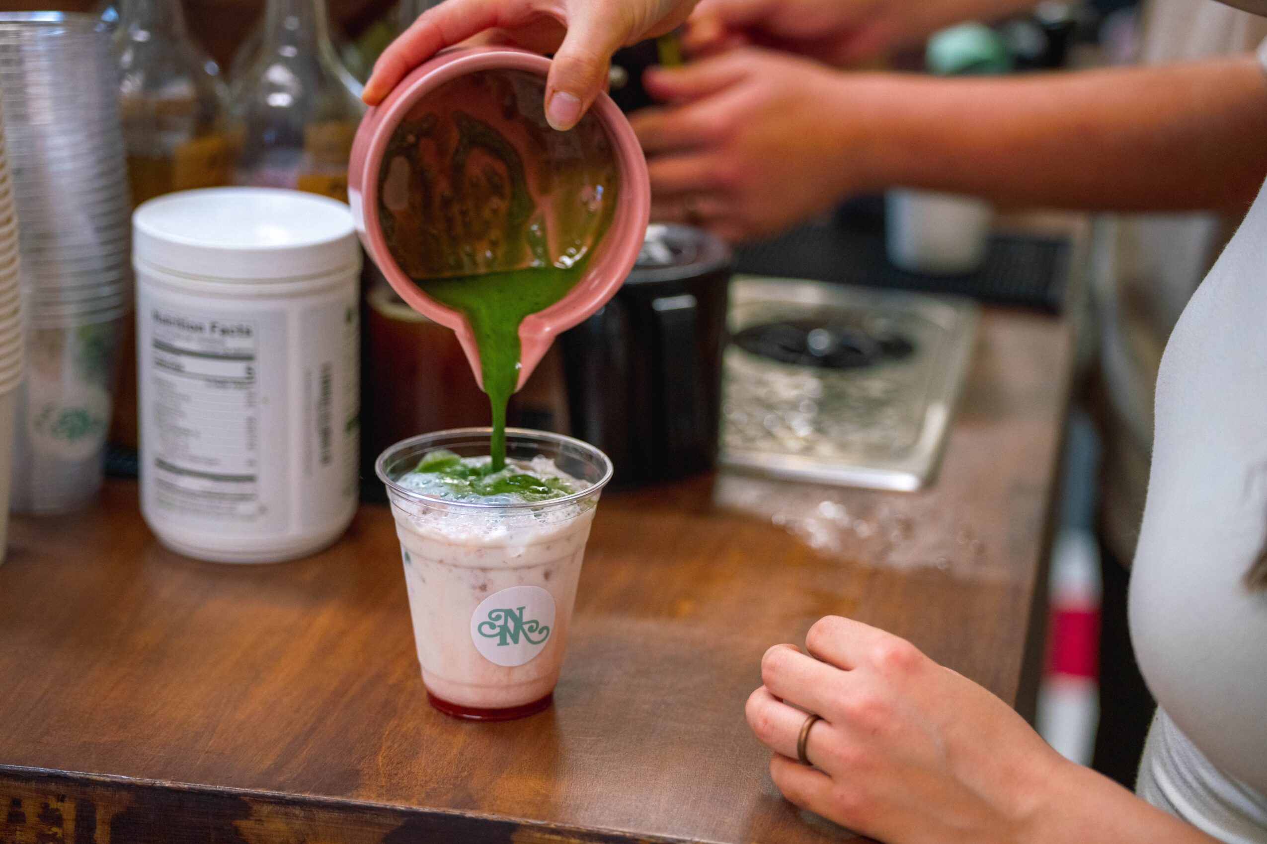 Person pouring green liquid into a cup on a wooden counter, with various containers and a stove in the background.