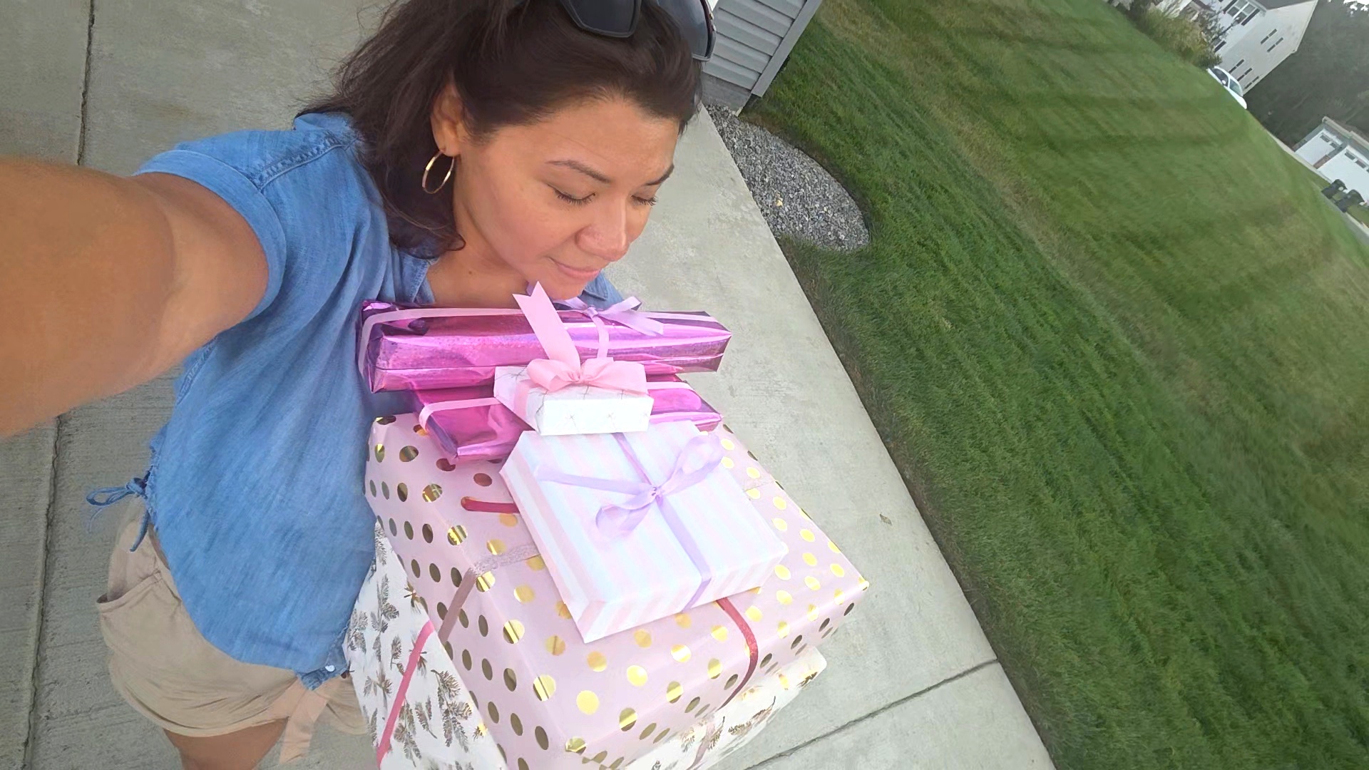 Woman taking a selfie outdoors with a large, wrapped gift box, grass, and sidewalk in the background.