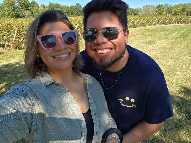 Two smiling people wearing sunglasses outdoors on a sunny day, with trees and grass in the background.
