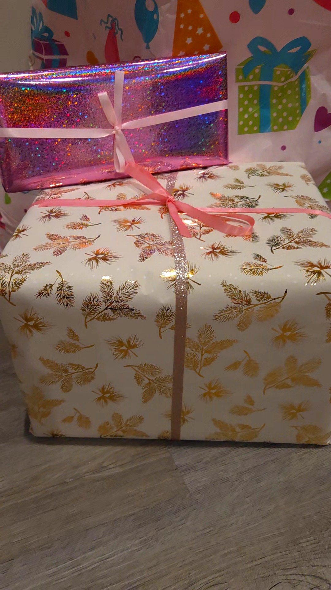 Three wrapped gift boxes stacked, with a background of colorful gift illustrations, one with pink holographic wrapping and ribbons.