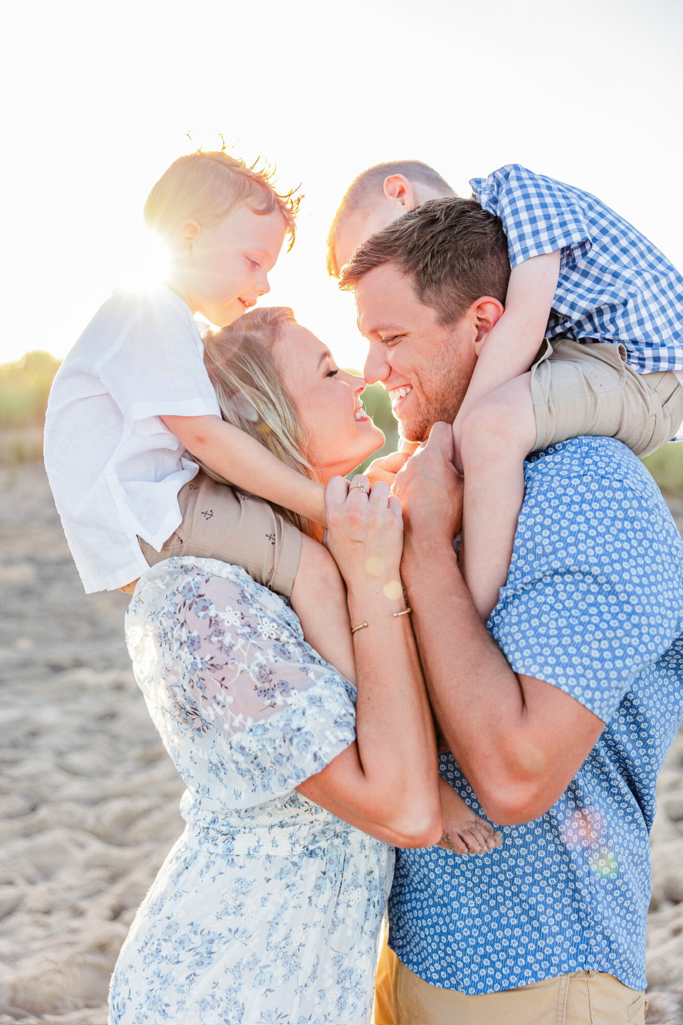 Family of four smiling and playing together outdoors, sunlight behind them, on a sandy surface.