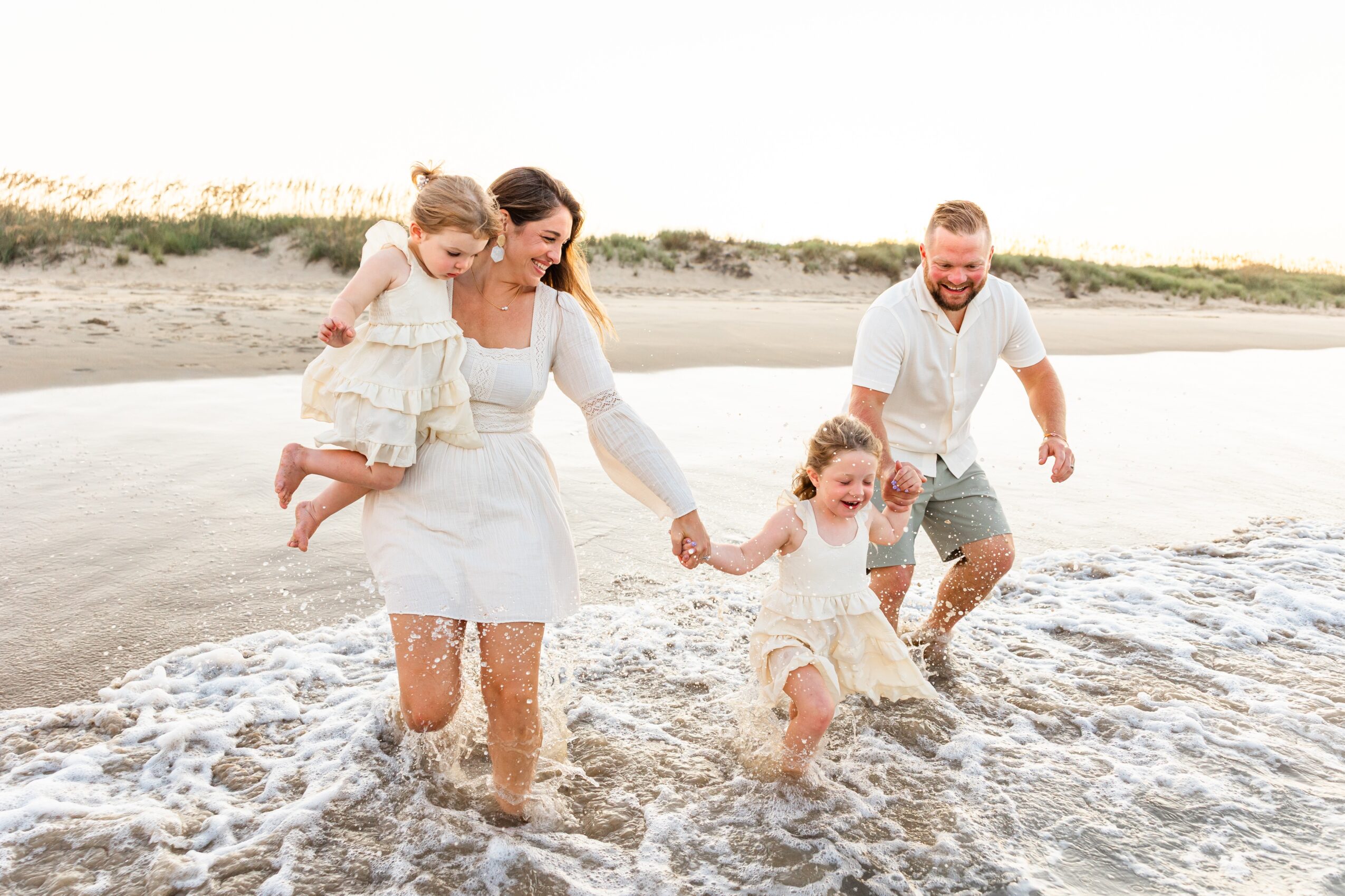 Family of four walking in shallow water on beach, holding hands, smiling, with sandy shoreline and sky in background.