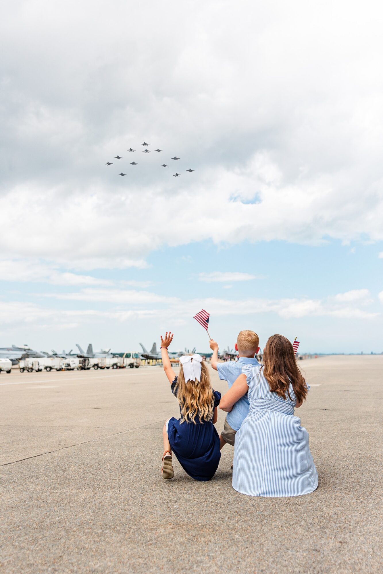 Two women and a girl sit on the ground watching a formation of planes fly overhead, with a cloudy sky and boats in the background.