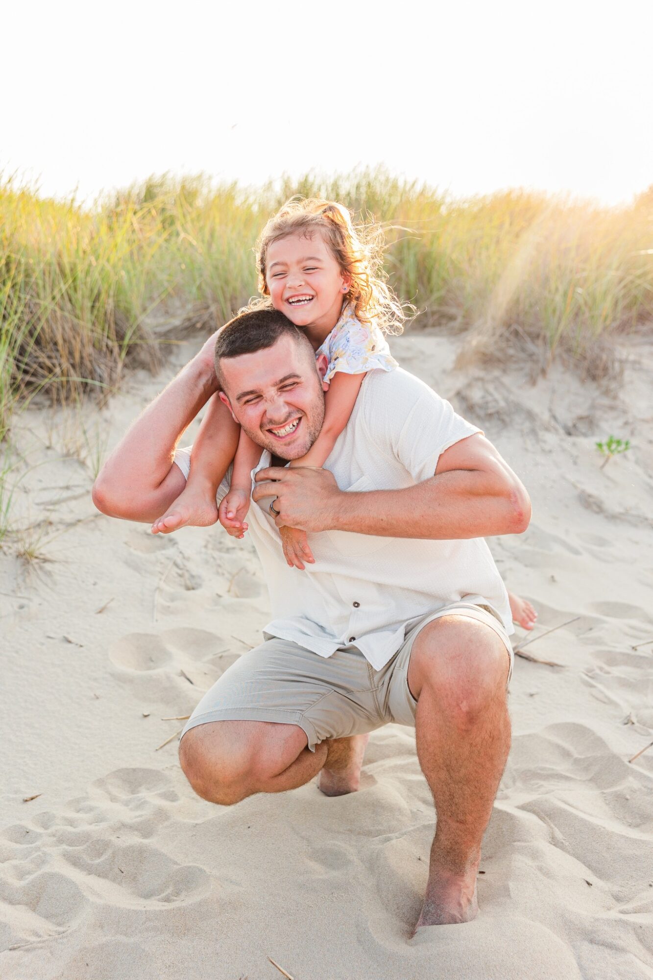 A man and a young girl are smiling and playing on a sandy beach with grass in the background.