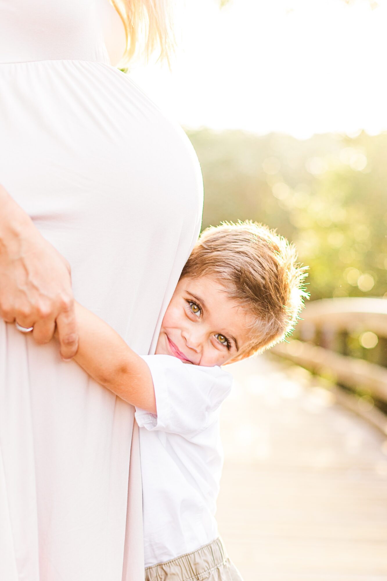Child hugging a pregnant woman's belly outdoors, smiling, with sunlight and greenery in background.