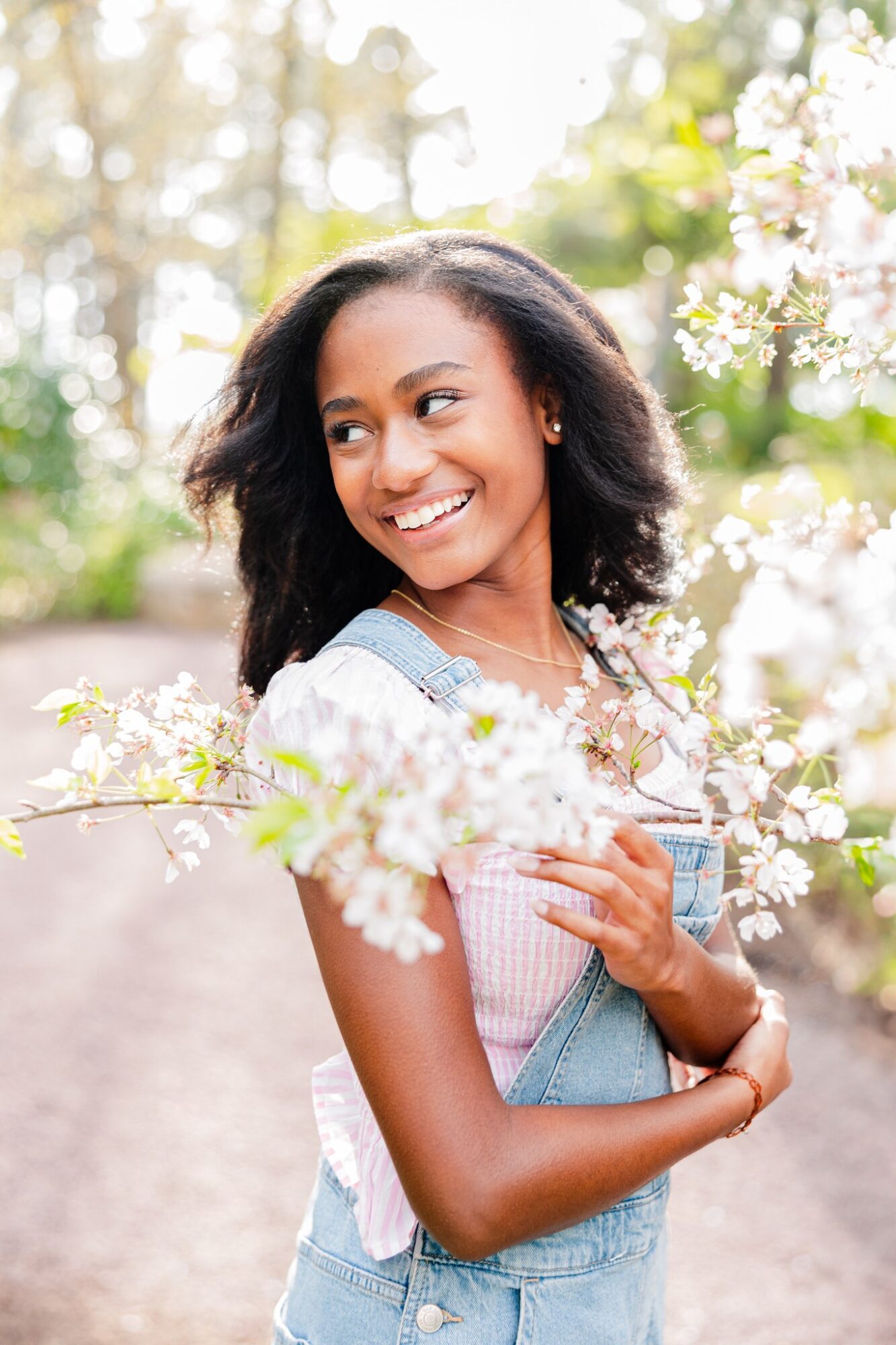 Smiling woman with curly hair outdoors among blooming flowers, wearing a white top and denim overalls.