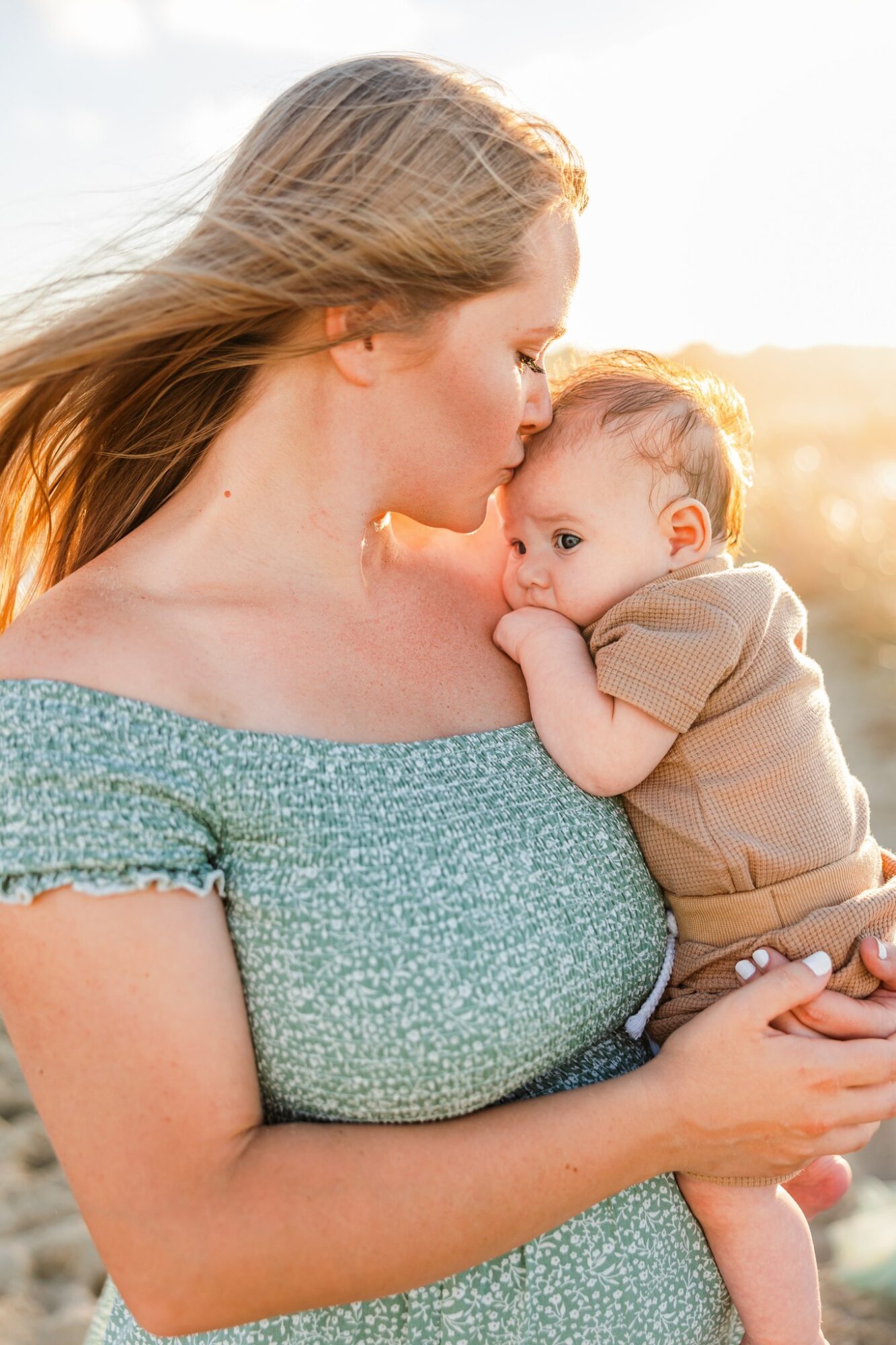 Woman holding a baby outdoors in sunlight, close-up of their faces, woman kissing baby's head, warm and tender scene.