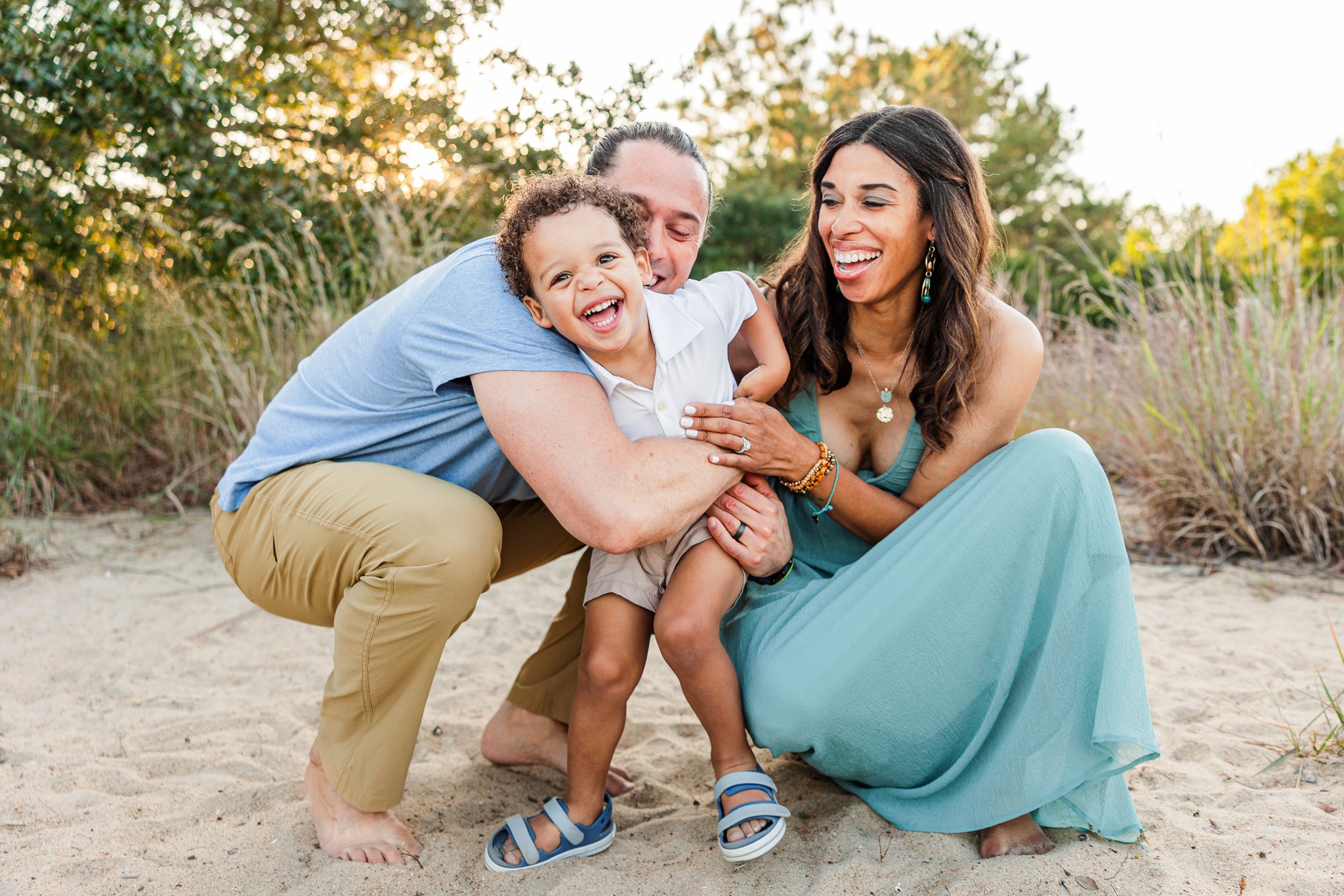 Family of three smiling and playing outdoors on sandy ground with trees in background.