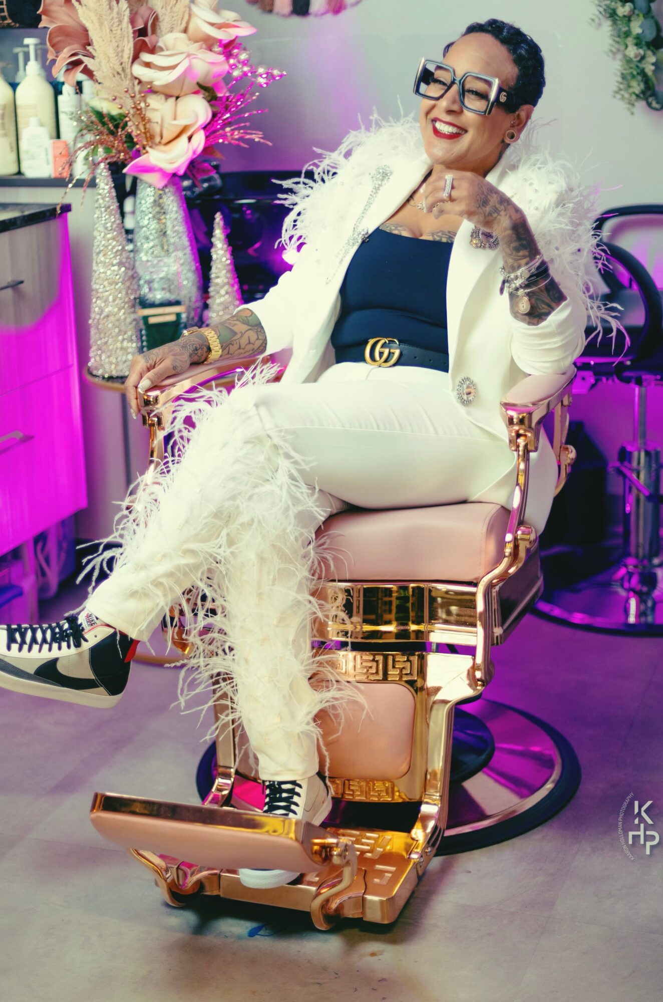 Woman with short dark hair and glasses, smiling, sitting in a luxurious chair with a feathered coat, in a decorated room.