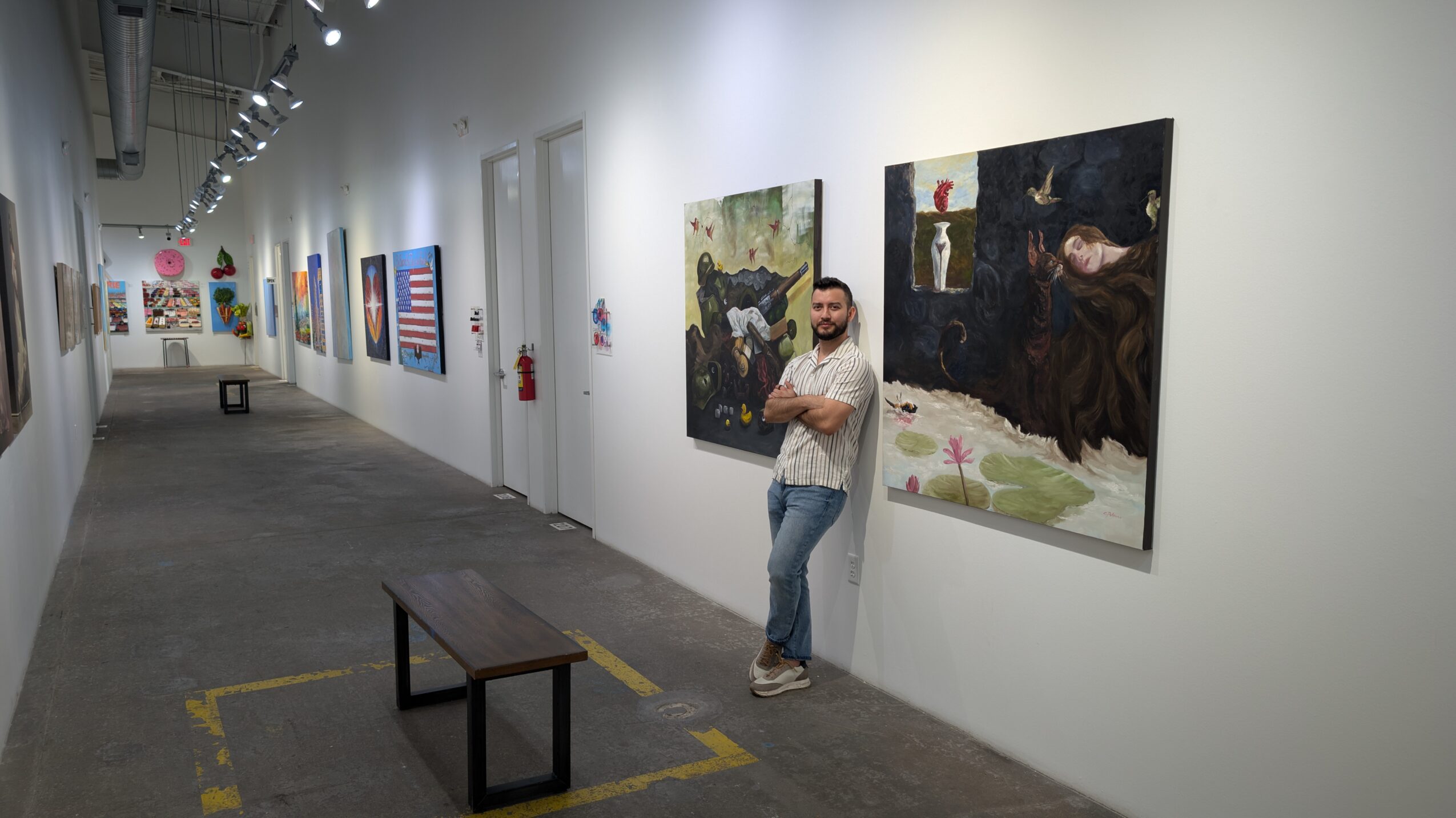 Man standing in art gallery with paintings on white walls, bench in foreground, gray floor, and ceiling lights. Word count: 27
