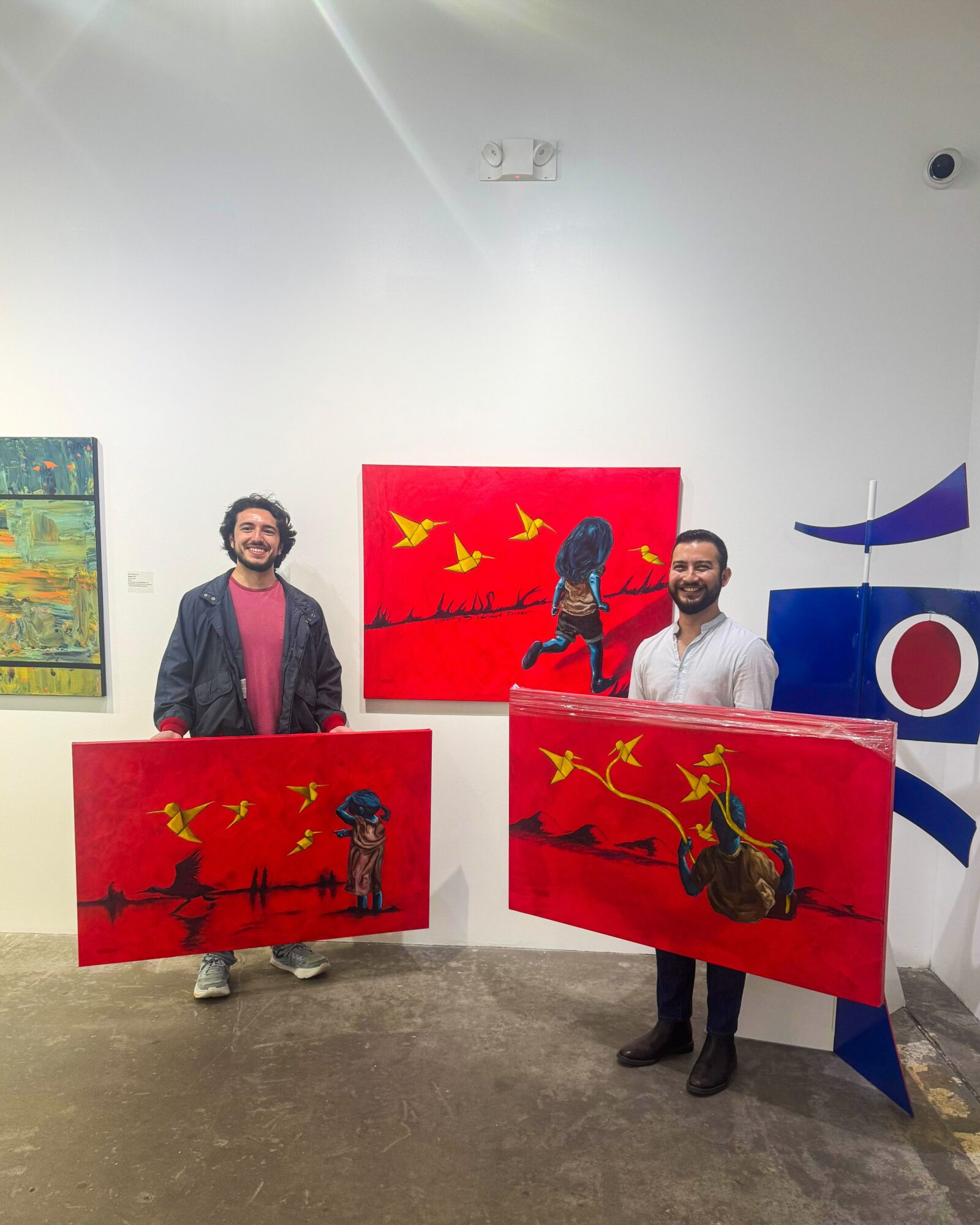 Two men stand in an art gallery holding red paintings with yellow birds and dark figures, white wall background.