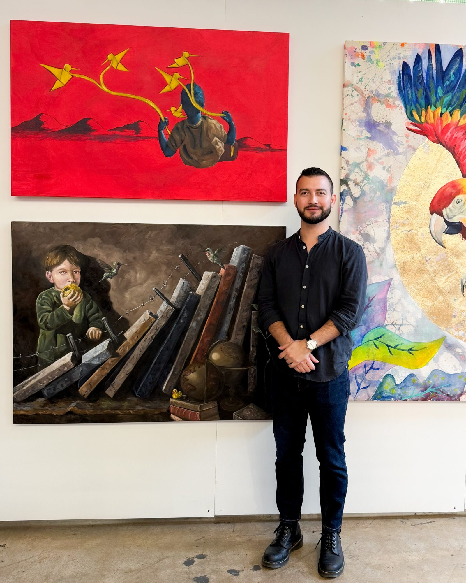 Man with beard and dark clothing standing in front of colorful paintings, including a red artwork with yellow birds and a vibrant parrot.