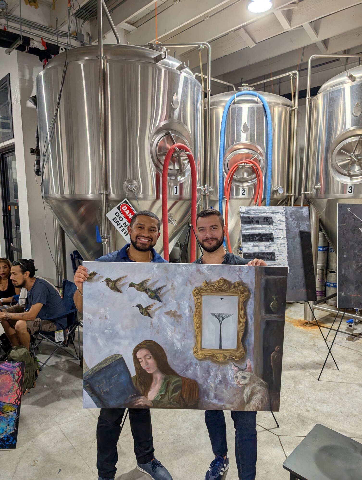 Two men holding a painting in a brewery with large stainless steel tanks in the background.