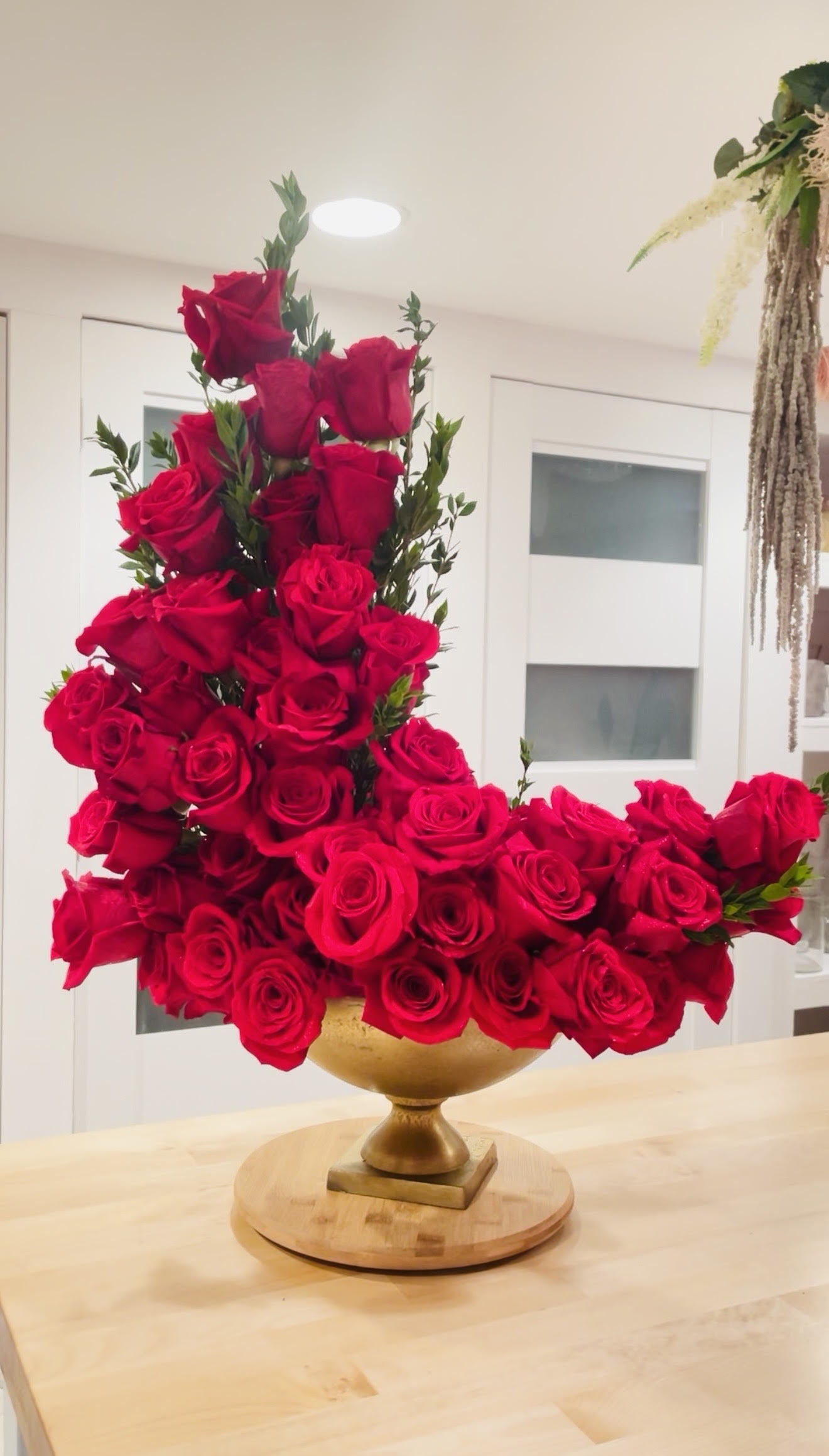 A large bouquet of red roses arranged in a gold vase on a wooden surface, with a white background and window.