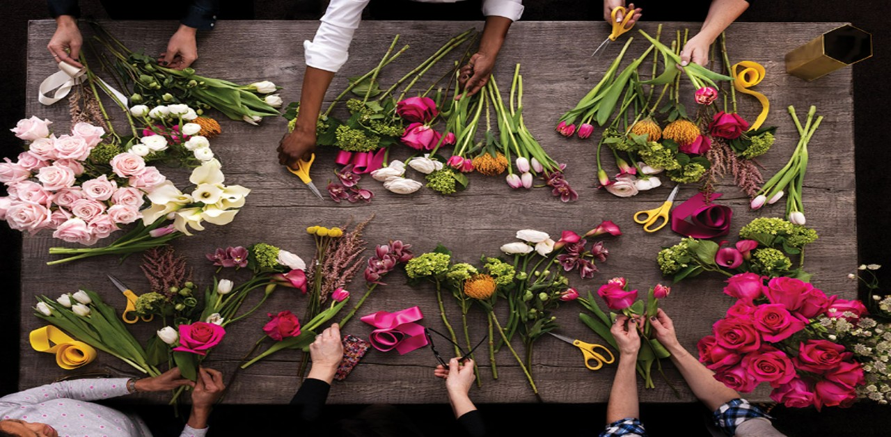 People arranging pink, yellow, and orange flowers on a dark table, with hands reaching in from all sides.