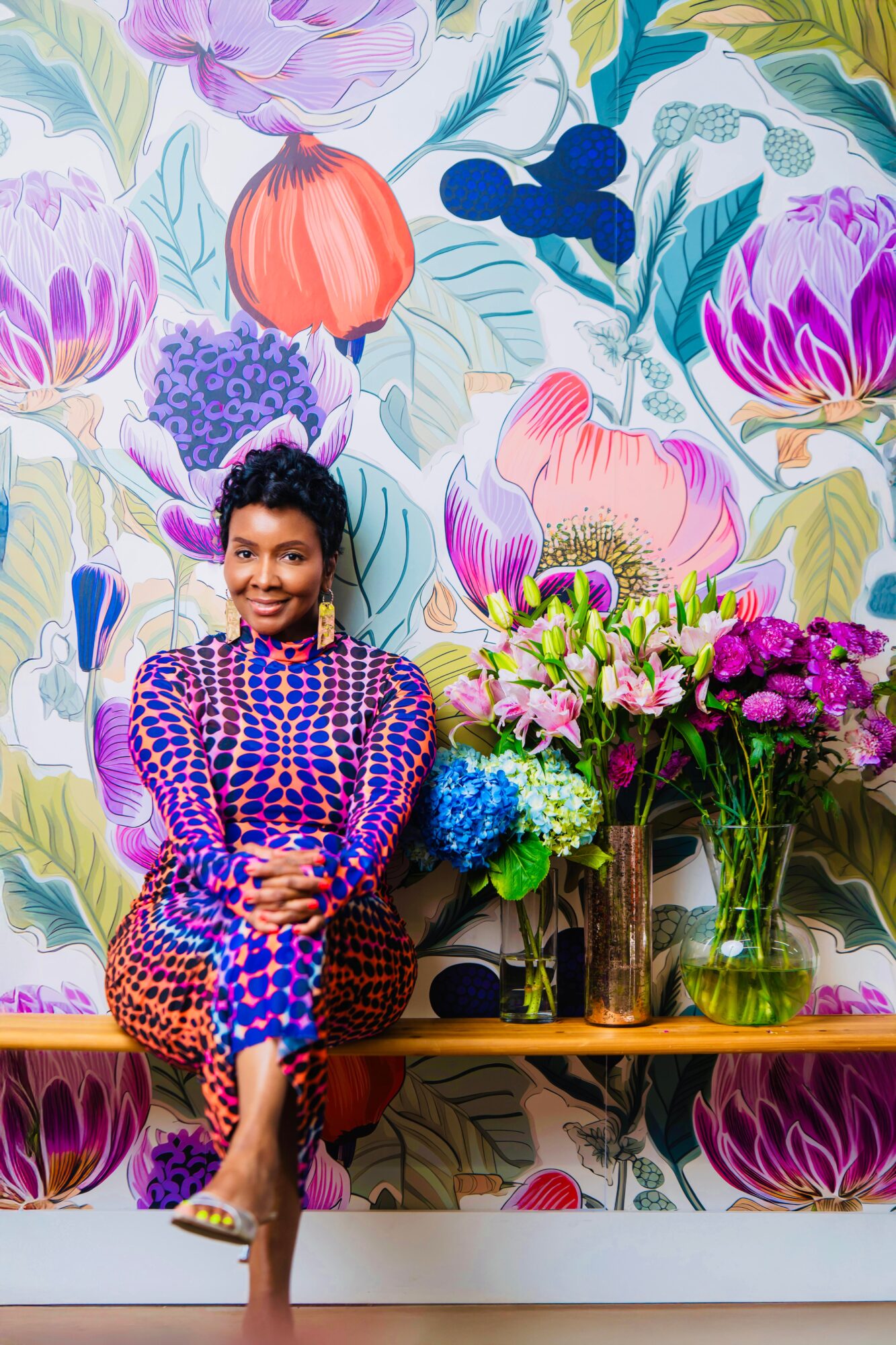 Woman sitting on a bench with colorful flowers and a floral wallpaper background, smiling, wearing a patterned dress.
