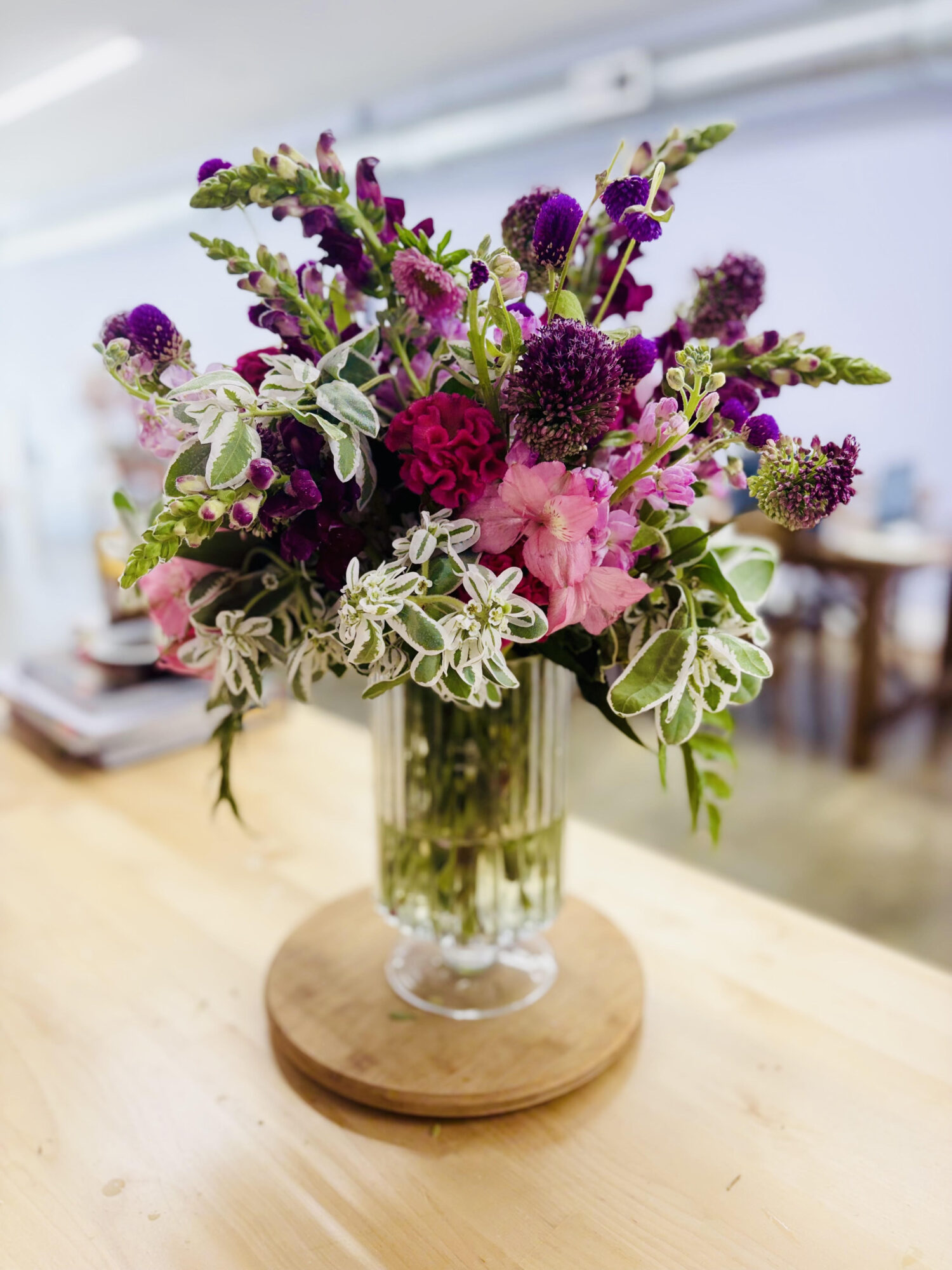 Vase with pink, purple, and green flowers on a wooden table, background of a room with chairs and a window.